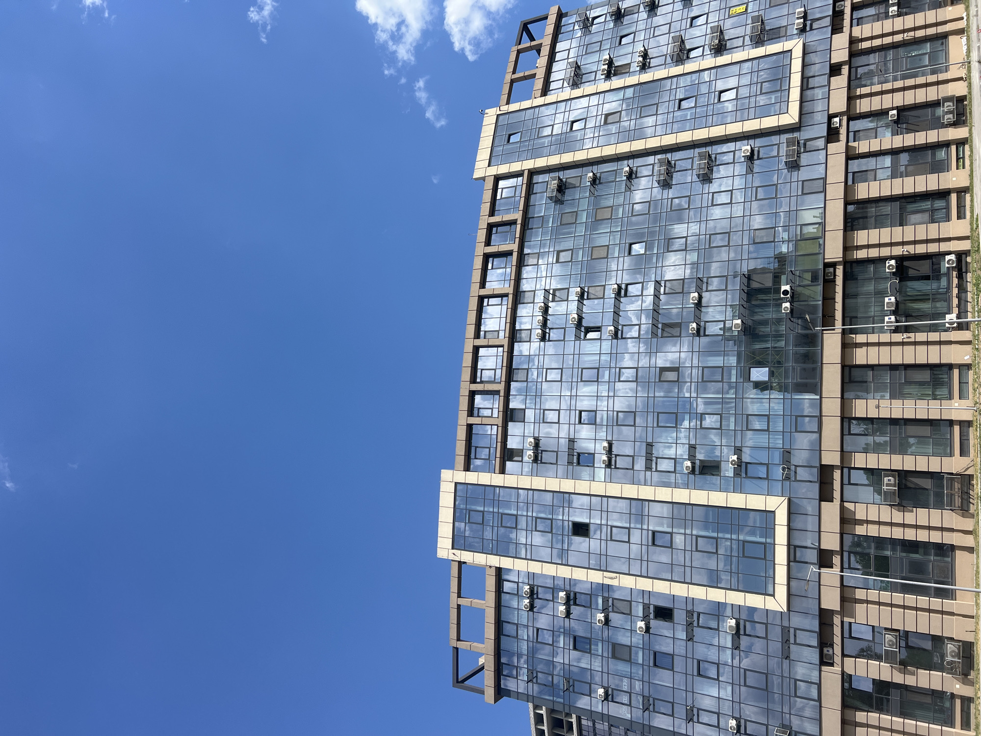 A low-angle, full-frame shot shows the exterior of a modern, multi-story building under a clear blue sky with scattered white clouds. The building features a curved facade predominantly clad in reflective blue-tinted glass windows, with vertical bands of beige stone or paneling accentuating its design. Numerous air conditioning units are visible mounted outside many of the windows across several floors. The lower section of the building has a more solid facade with recessed windows and vertical accent lines. A street light is positioned in front of the building. The scene appears to be in an urban environment, though no specific landmarks or identifying features are present in the provided image.