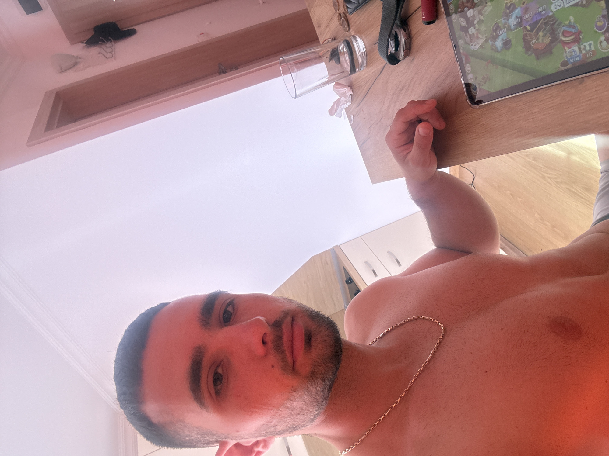 A man with short dark hair and a beard is seen from the chest up, facing the camera. He is shirtless and wearing a gold chain necklace. The man is seated at a wooden table, and on the table, there is a glass of water, a watch, and a tablet displaying a game. In the background, there are doors and a wall. The scene appears to be an indoor setting, likely a residence, in Nesebar, Bulgaria.