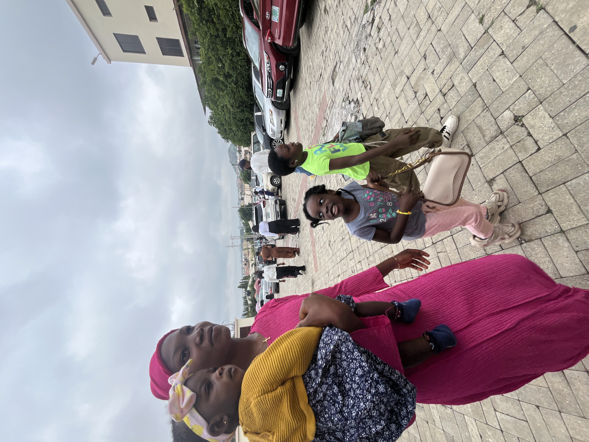 A group of people, including a mother carrying a baby, walk across a paved outdoor area near parked cars in Abuja, Nigeria. The mother, wearing a pink headscarf and dress, holds a baby in her arms. A younger girl in a green t-shirt and khaki pants walks beside her, and another girl in a grey t-shirt and pink pants walks slightly ahead. In the background, several cars are parked, and a building with multiple windows is visible. The sky is overcast.