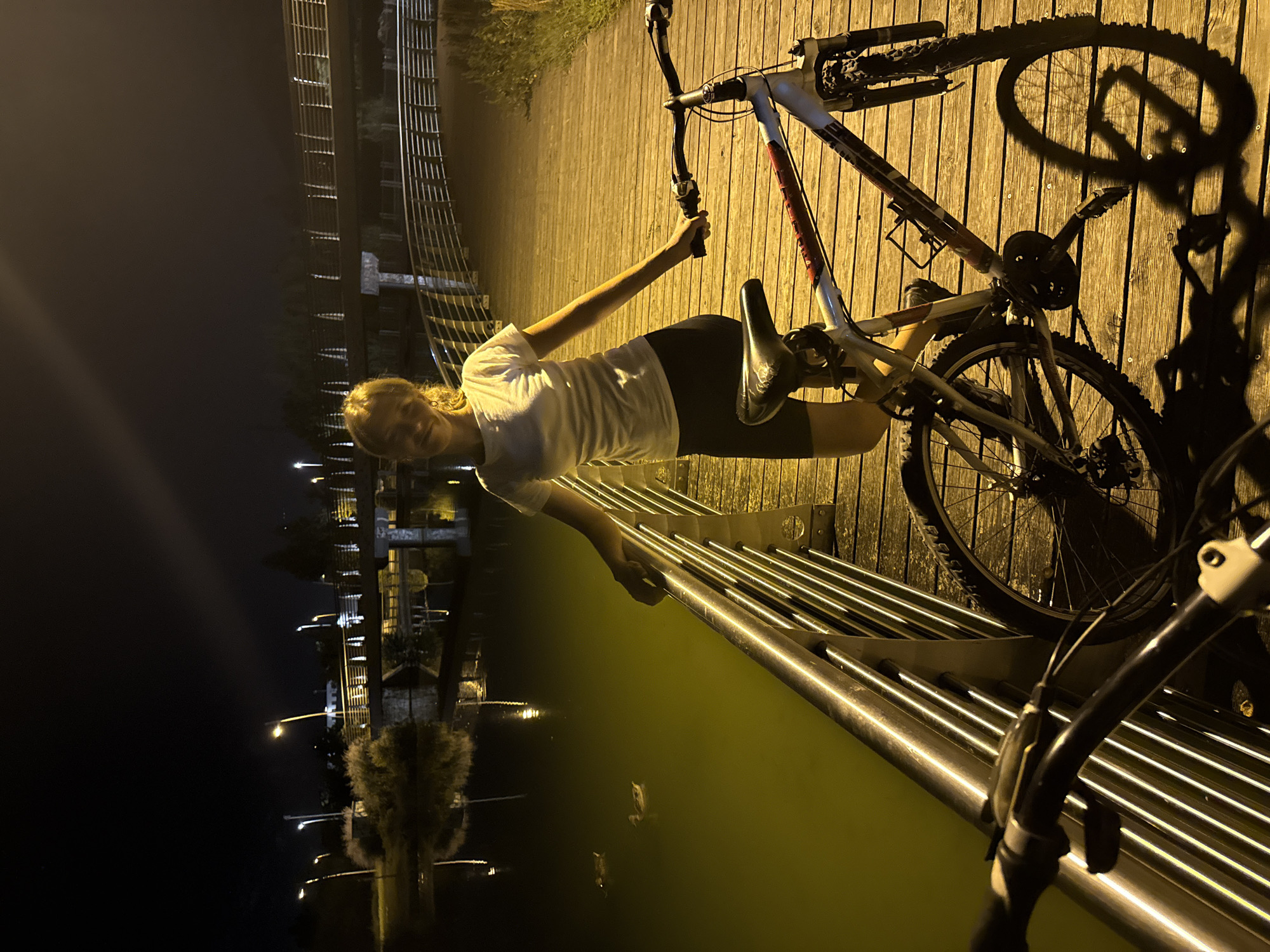 A young woman stands on a wooden boardwalk at night, next to two bicycles. She is wearing a white t-shirt and dark shorts, and her blonde hair is pulled back. She is smiling and looking at the camera. One bicycle is in the foreground, and another is partially visible behind it. The boardwalk overlooks a body of water, where several ducks are swimming. In the background, there is a bridge illuminated by streetlights, and the city skyline of Trento, Italy, is visible. The scene is lit by artificial lights from the streetlamps and the bridge.
