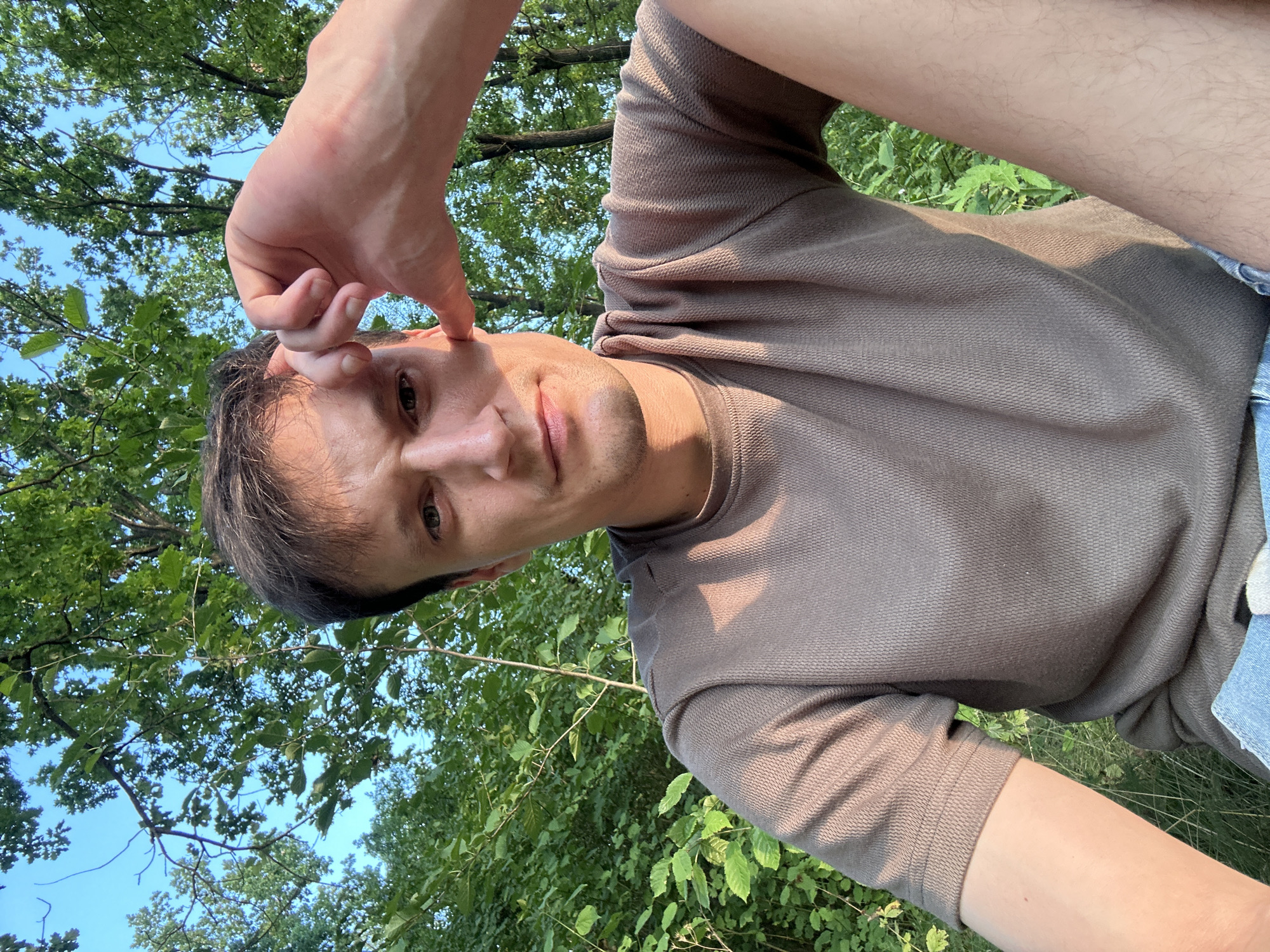 A young adult male is taking a selfie outdoors with his right hand partially covering his face. He is wearing a brown t-shirt and blue denim shorts. The background consists of lush green foliage and trees, suggesting a natural setting, possibly a park or forest. The lighting indicates it is daytime, with sunlight filtering through the leaves. The location is specified as Makiv, Ukraine.