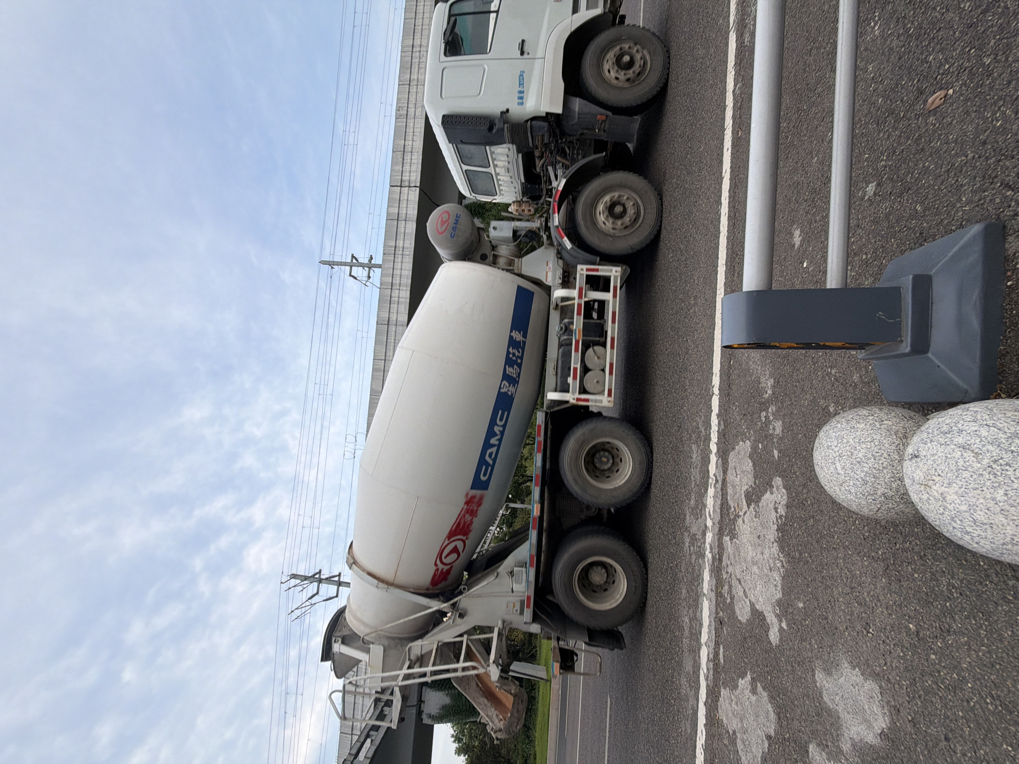 A white CAMC cement mixer truck is parked on a paved road in Jiangjin, China, with an overpass and electrical wires visible in the background under a cloudy sky. The truck's large cylindrical mixing drum is prominently featured, with the CAMC logo visible in red and blue. The truck has dual rear wheels on each side. In the foreground, two large, rounded, granite-like bollards and a metal barrier are situated on the roadside. The road surface shows some wet patches. There are no people visible in the image.
