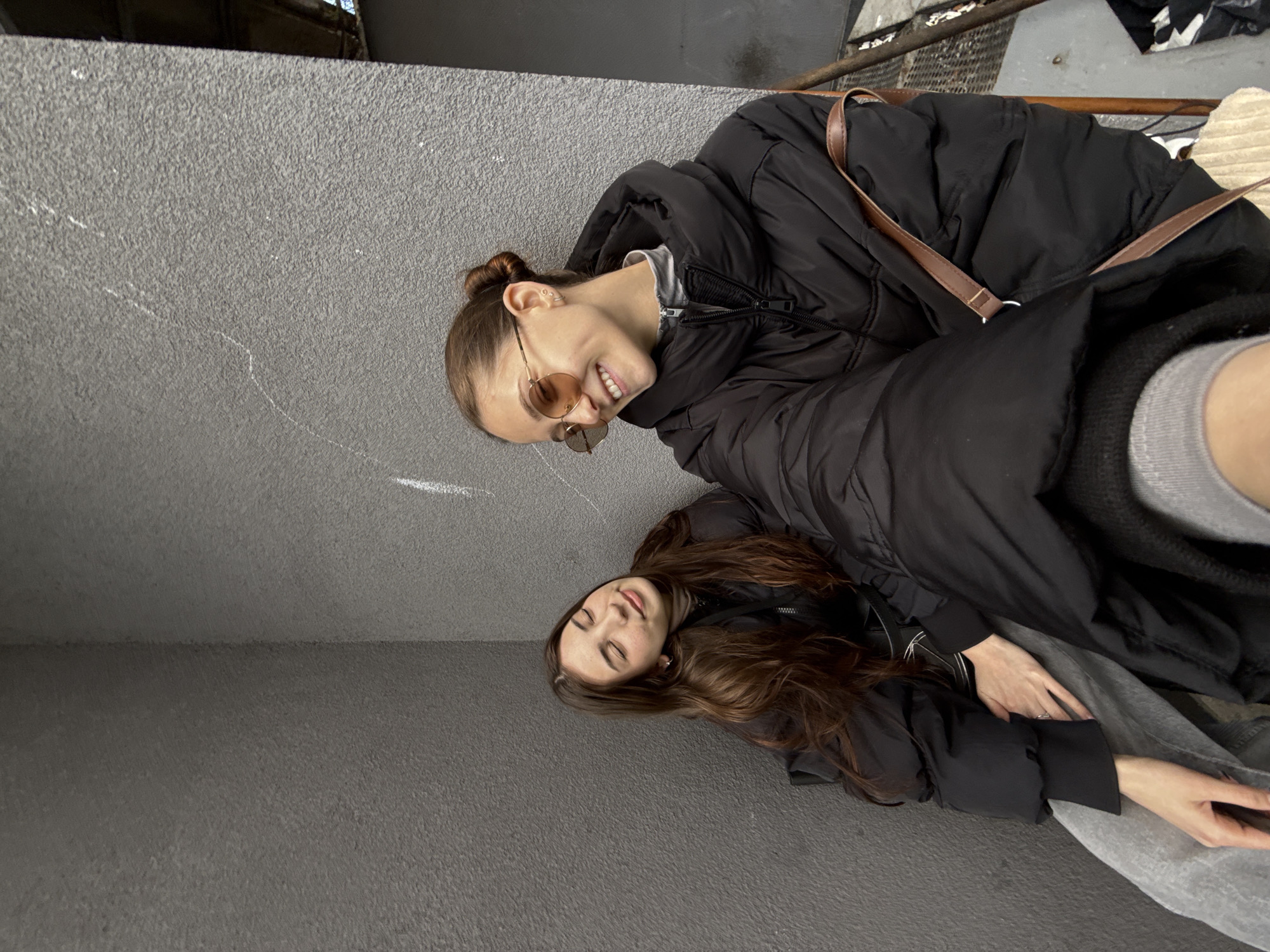 Two women are sitting against a concrete wall. The woman in the foreground is smiling, wearing rose-tinted glasses, a black puffy jacket, and a brown purse. The woman in the background is wearing a black jacket and gray pants. The location appears to be an urban area with concrete structures, potentially in Sofia, Bulgaria.