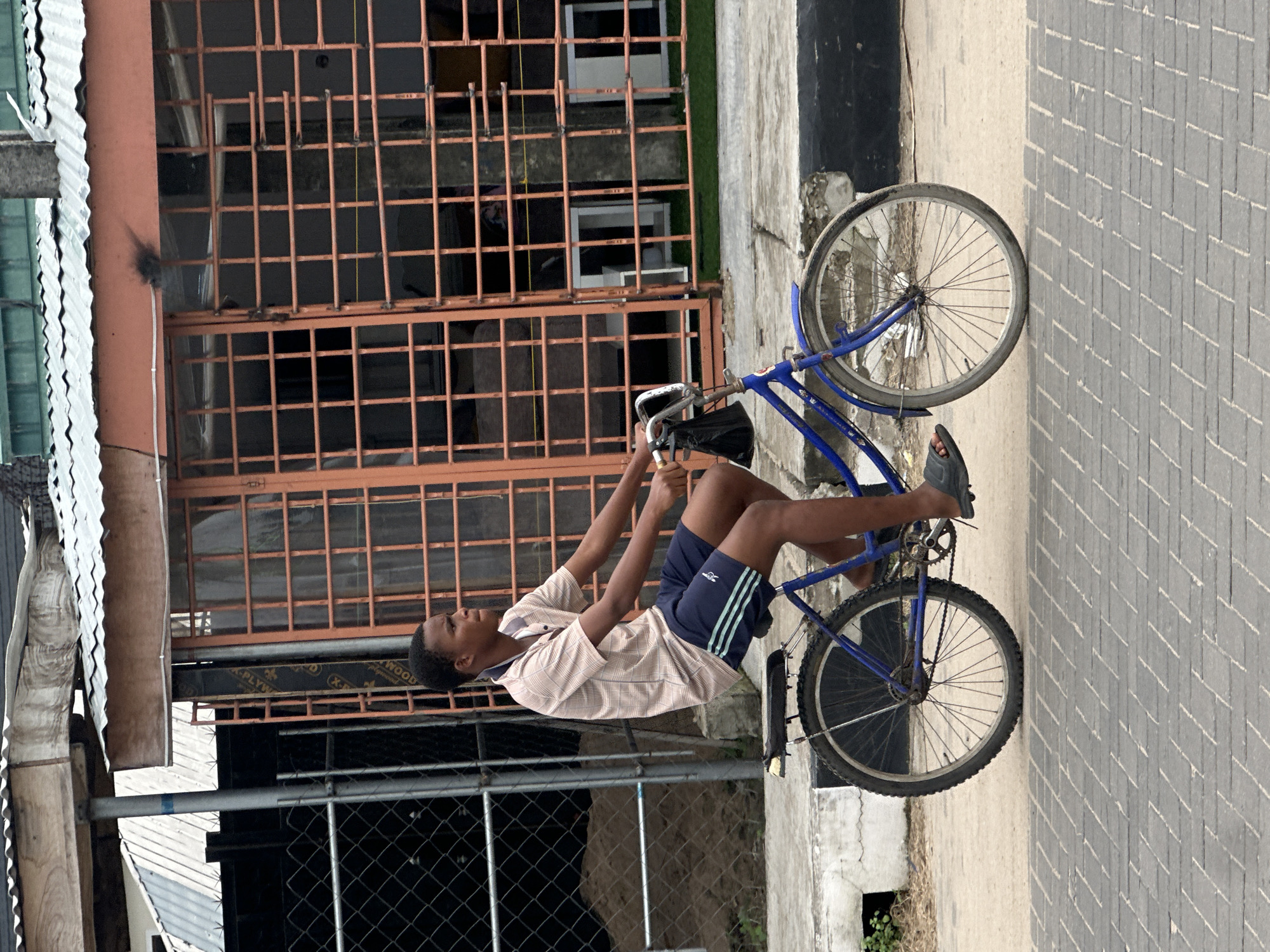 A young male rides a blue bicycle on a paved road in Badagry, Nigeria. The man wears a light brown plaid short-sleeved shirt and dark blue shorts with light blue stripes. He has black hair and is wearing black sandals. He is looking to his right. The bicycle has a black bag hanging from the handlebars. The background features a building with an orange-barred facade and a corrugated metal roof, with a chain-link fence to the left. The road is paved with grey bricks.