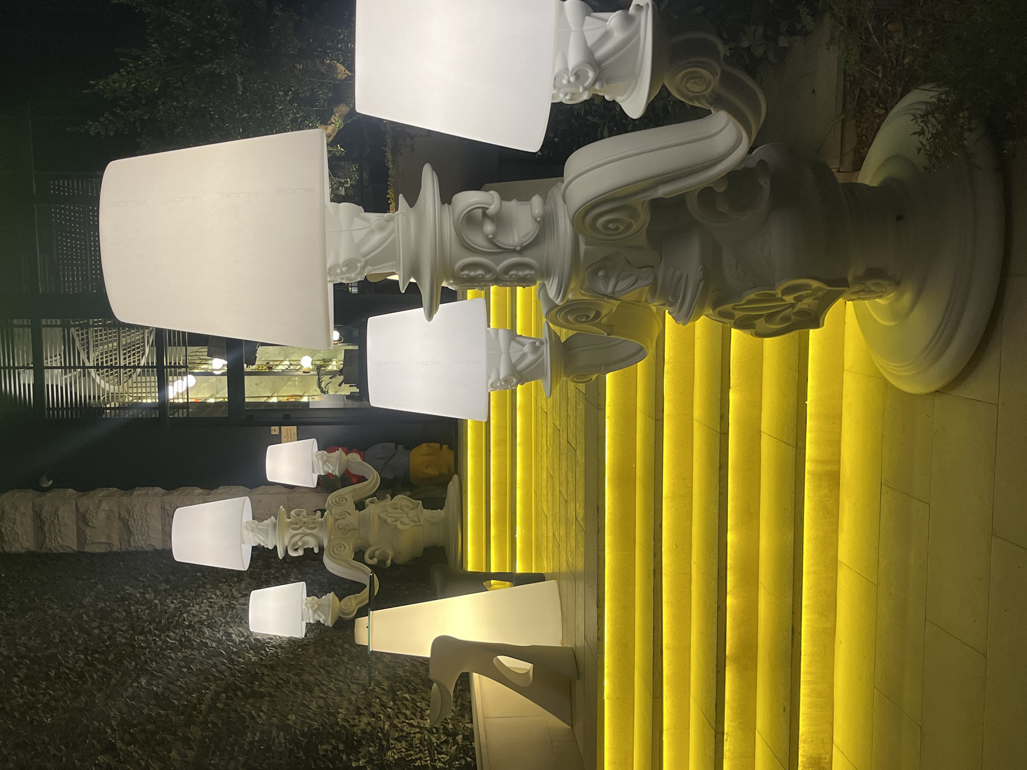 The image shows a row of ornate, white lamps with white conical lampshades lining a set of brightly lit yellow stairs. The scene appears to be outdoors at night. The base of each lamp features baroque-style carvings. The steps are bathed in a soft, glowing yellow light, creating a striking contrast. In the background, on the left, there are trees and a building with visible windows. Further back is another lamp, some of which are partially obscured. The location is Budva, Montenegro.