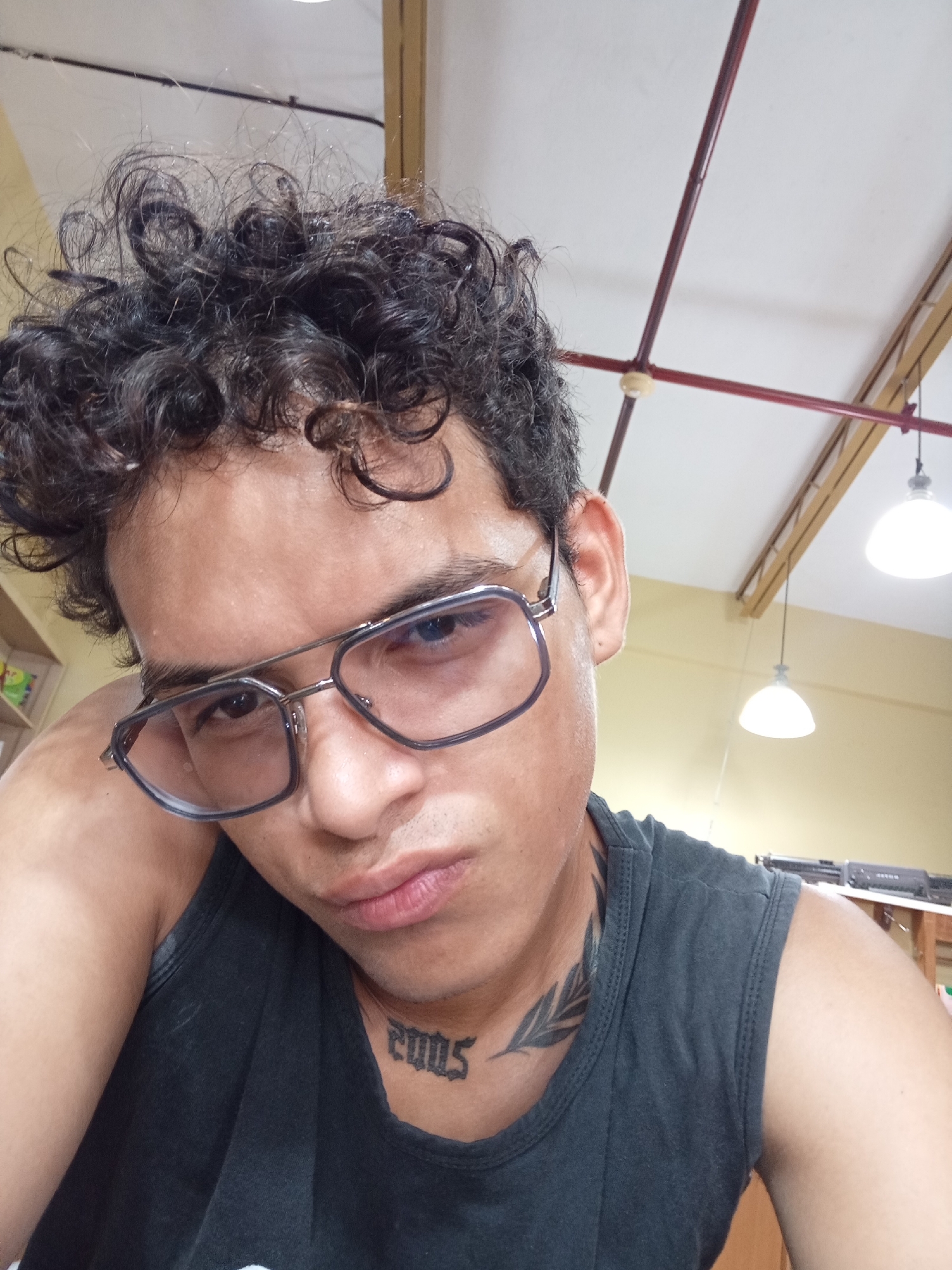 The image depicts a young adult, possibly male, with medium-dark skin and dark, curly hair, situated indoors in Juan Griego, Venezuela. The individual wears metal-framed glasses with dark-rimmed lenses and a dark grey or black sleeveless tank top. Visible tattoos include the year "2005" in stylized script on the front of the neck and a dark leaf or branch design on the left side of the neck. The subject is posed with their left forearm and hand resting against their head, looking directly towards the viewer with a neutral or slightly pensive expression. The background features a white ceiling with visible reddish-brown structural beams or pipes and light yellow walls. Multiple illuminated white dome-shaped pendant lights hang from the ceiling. Blurred elements in the background suggest shelving or furniture, with what appears to be equipment visible on the right side.