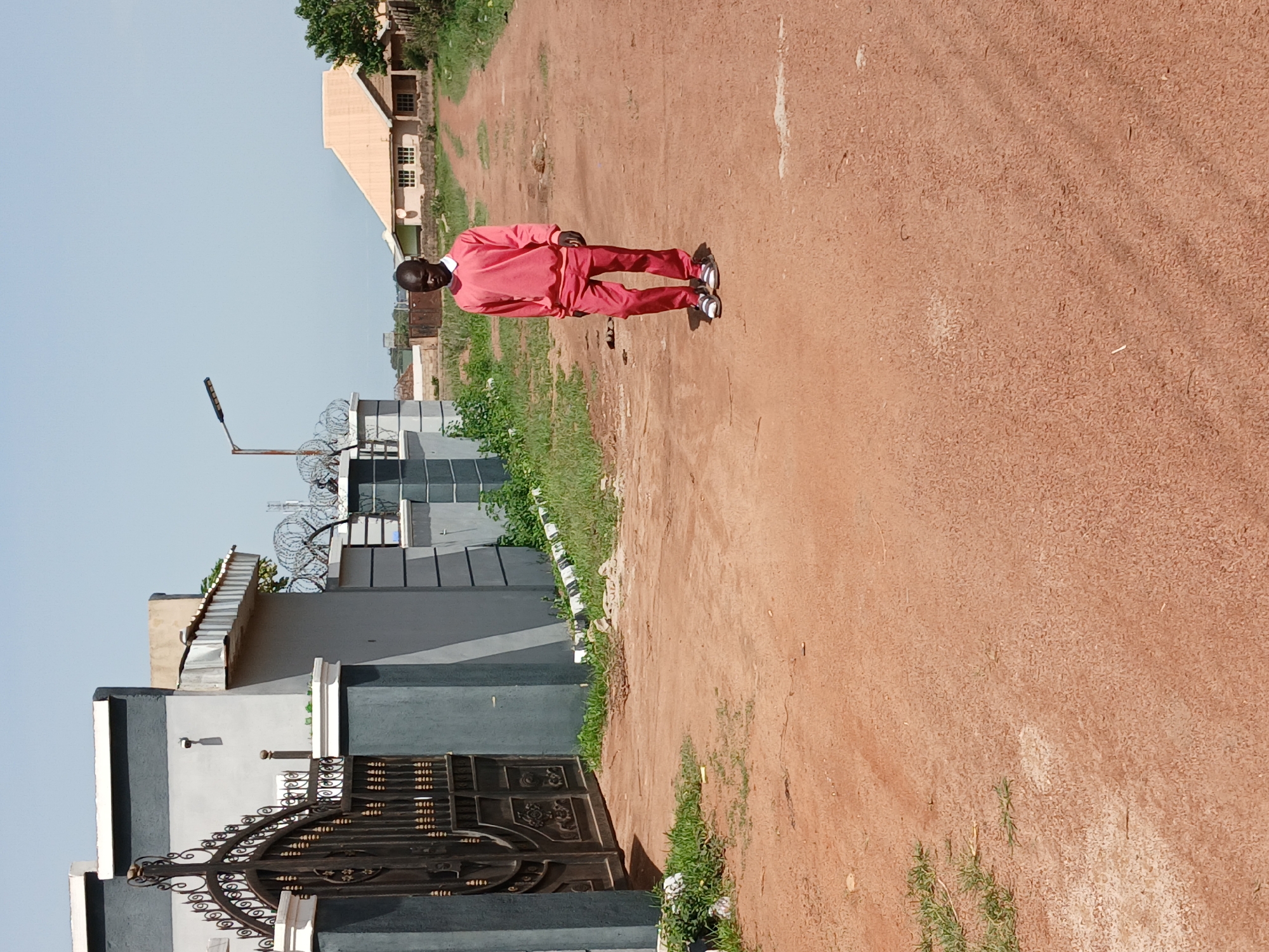 A young Black male wearing a coral-colored tracksuit and white and grey sandals walks away from a large, ornate black metal gate. The gate is part of a grey wall with white trim. Behind the gate is a building with a flat roof and grey walls, and some barbed wire on top of the wall. The man is walking down a reddish-brown dirt road that curves to the right. On the right side of the road, there is a light brown house with a light yellow roof and a fence. In the background, there are some trees and other buildings, including what appears to be a street lamp. The sky is clear and blue. The scene is set in Kwang, Nigeria.