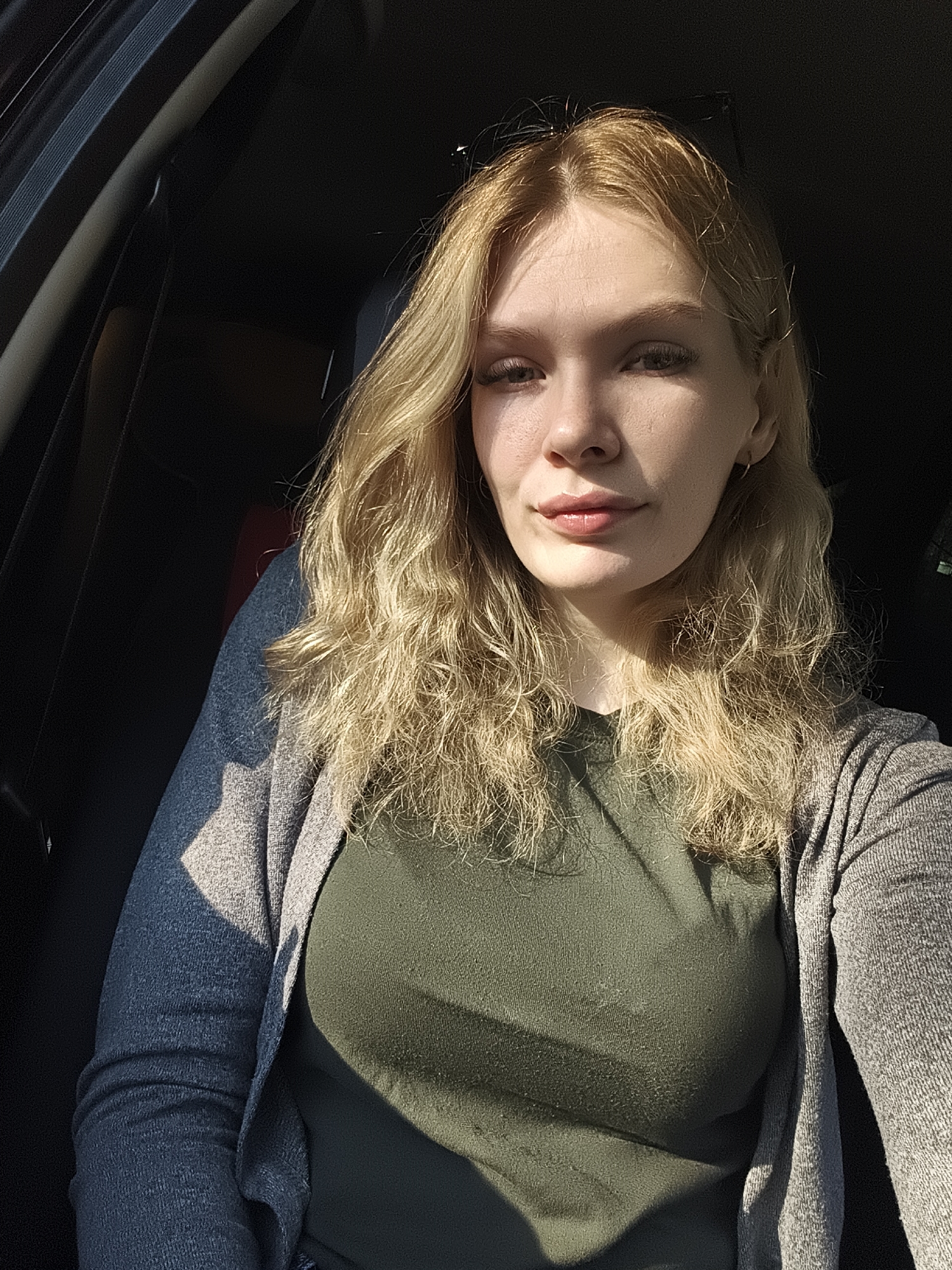 A young woman is photographed inside a car, seemingly in the driver's seat. She has blonde hair and is wearing an olive green t-shirt and a gray cardigan. The background is dark, likely the car's interior. There is nothing visible that would indicate the photo was taken in Izhevsk, Russia. There are no identifiable objects or interactions occurring. The environment appears to be a car interior with strong sunlight coming in from one side.