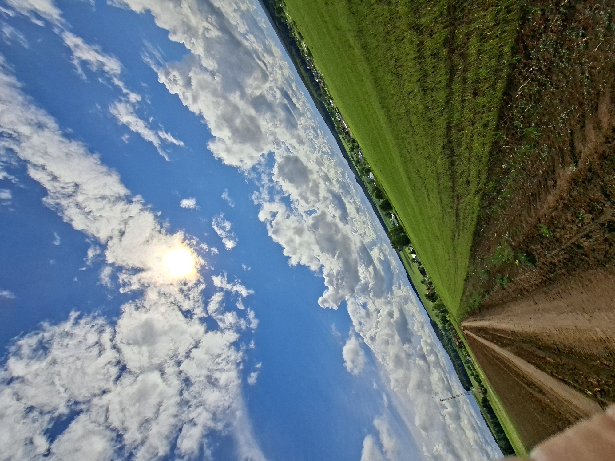 This image captures a vibrant rural landscape under a bright, partly cloudy sky. The scene is dominated by a clear blue sky, interspersed with numerous white, fluffy cumulus clouds. The sun, appearing bright and slightly hazy through a thin cloud, is visible in the upper left portion of the sky.
Below the expansive sky, the landscape unfolds with a wide, light brown dirt path or track prominent on the right side, leading into the distance. To the left of this path lies a vast, lush green field, indicating fertile or cultivated land. Further in the background, a line of trees or shrubs borders the field, behind which small buildings or a distant village can be discerned. A tall, thin structure, possibly a telecommunications tower, also stands faintly in the distant mid-ground.
The photograph depicts a daytime scene, likely mid-day given the high position and brightness of the sun, with fair weather and scattered clouds. A small portion of a hand or finger is visible in the very bottom right corner, suggesting the photo was taken handheld. There is no visible text in the image. The provided location context for this scene is Karsha, Russia.