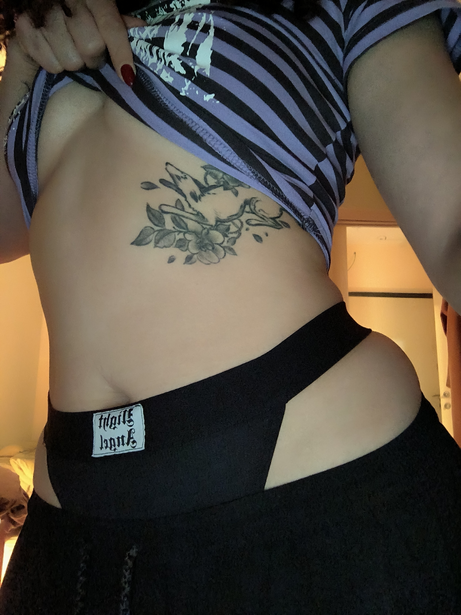A woman is posing for a close-up photograph indoors, lifting her striped shirt to reveal a tattoo on her ribcage depicting birds and flowers. She is wearing black pants with a unique cut-out design at the hip, displaying a white label on the waistband. The environment appears to be dimly lit, with a glimpse of a room and a doorway visible in the background. There is no information to indicate that the scene is located in Saint Petersburg, Russia.