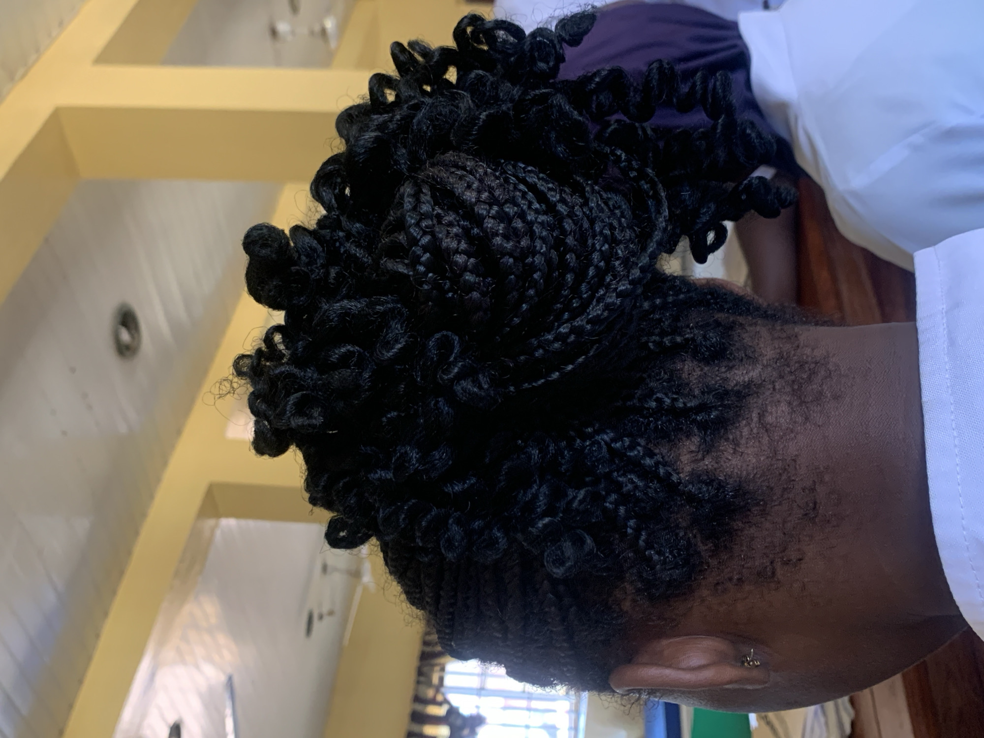 A close-up, eye-level indoor shot displays a person with elaborate braided hair. The hair is styled in dense braids with curled ends. A portion of the person's dark skin and ear are visible. They are wearing a white garment. The background shows a yellow wall or structure. The location is potentially within the city of Apata Yakuba, Nigeria.