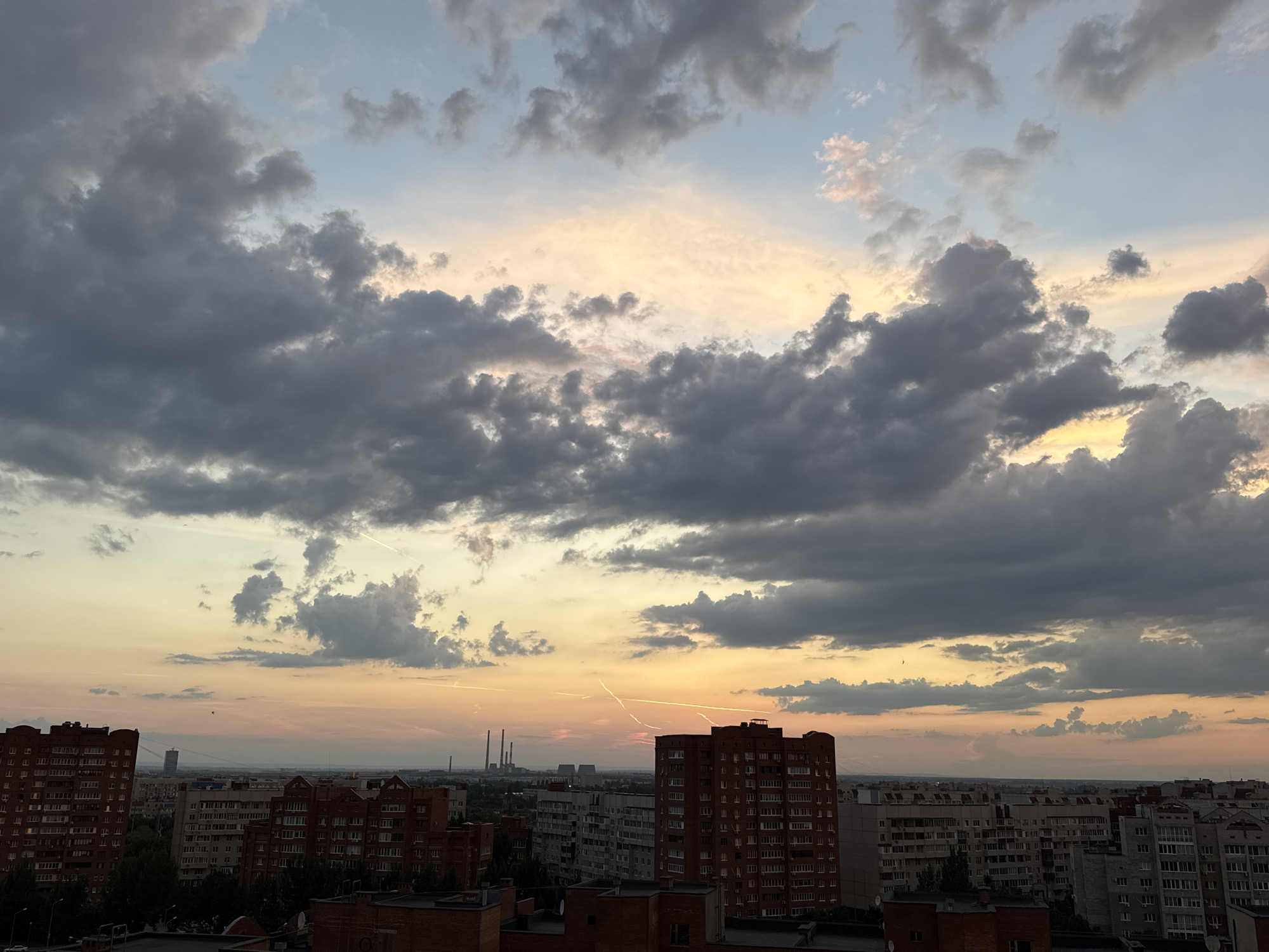 The image depicts a cityscape at dusk, characterized by a dramatic sky filled with a mix of dark and light clouds. The setting sun casts a warm, orange and pink glow across the horizon, contrasting with the cooler blues and grays of the upper sky. Several buildings, predominantly apartment blocks, are visible in the mid-ground and foreground. These buildings appear to be of various heights and architectural styles, with some featuring reddish-brown brick facades and others a lighter gray concrete. In the far distance, a cluster of industrial structures, possibly a power plant with tall smokestacks, is discernible. The overall scene suggests an urban environment during the transitional light of evening.