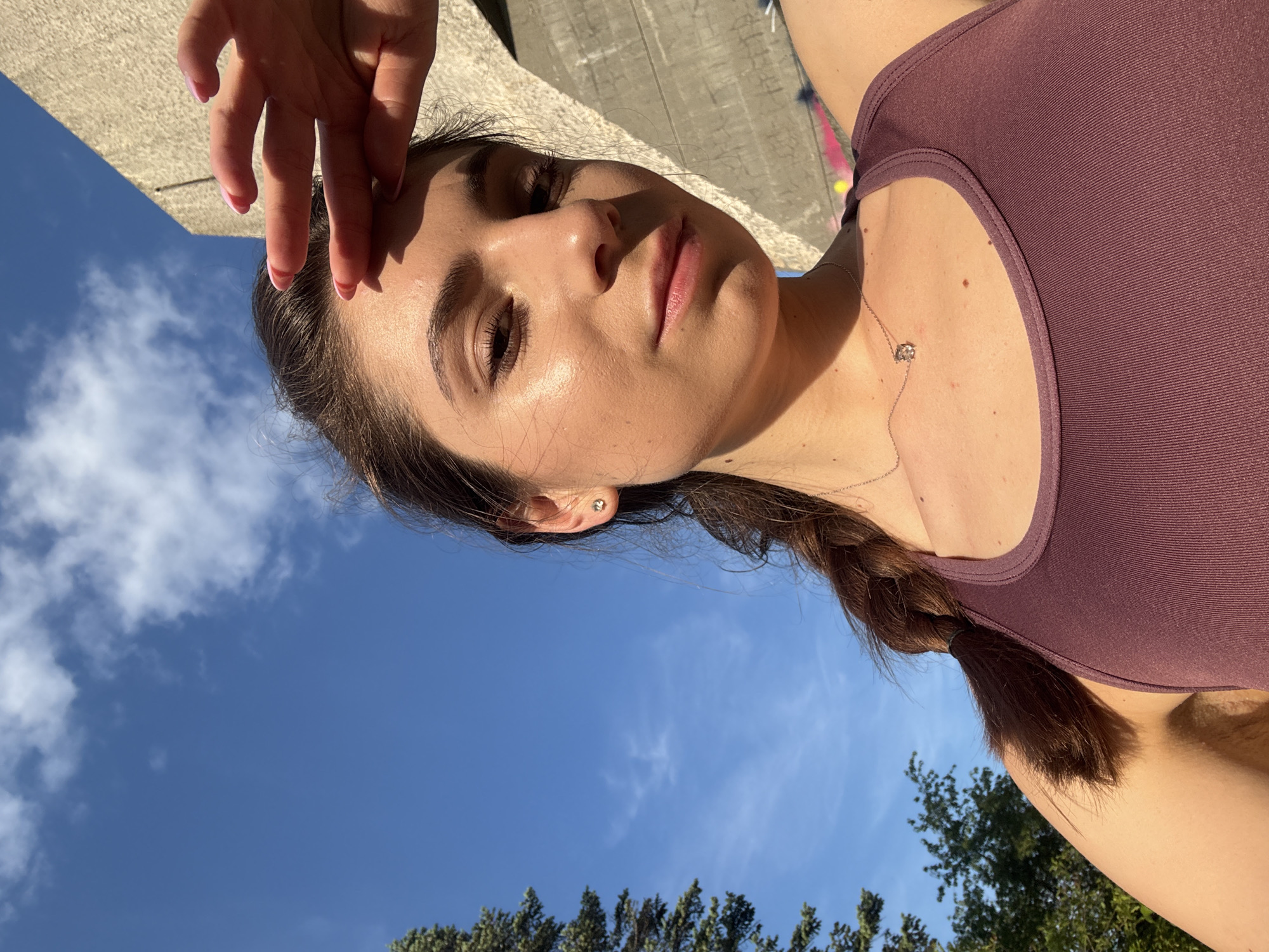 A young woman is captured in a close-up, self-portrait taken outdoors on a sunny day. She is wearing a purple athletic top and a delicate silver necklace with a small pendant. Her dark hair is styled in a braid that rests on her shoulder. Her right hand is raised to her forehead, partially obscuring her brow and casting a shadow on her face. The lighting suggests it is daytime, likely late afternoon, with the sun casting warm highlights on her skin.
The background is a split scene: the upper portion shows a textured concrete surface, possibly a wall or a building, with some graffiti visible. The lower portion of the image reveals a clear blue sky with scattered white clouds, and the tops of green trees are visible at the bottom edge. The perspective is as if the photo is taken from a low angle looking upwards. The overall mood is relaxed and casual, hinting at an outdoor activity or a moment of leisure. There is no visible text in the image. The location context provided is Varna, Bulgaria.