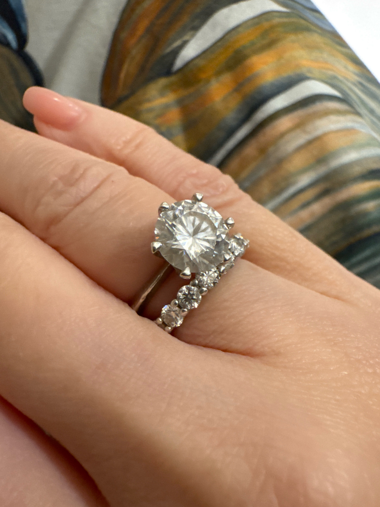 A close-up, overhead shot of a hand wearing two diamond rings is shown. The hand is lightly tanned, and the fingernails are painted a light pink. The left ring features a large, round-cut diamond as the centerpiece, with smaller diamonds accenting the band. The right ring is a delicate band encrusted with small diamonds. The background is blurred, but appears to be a fabric with abstract patterns in shades of brown, orange, and blue. The image was taken at an undisclosed location.