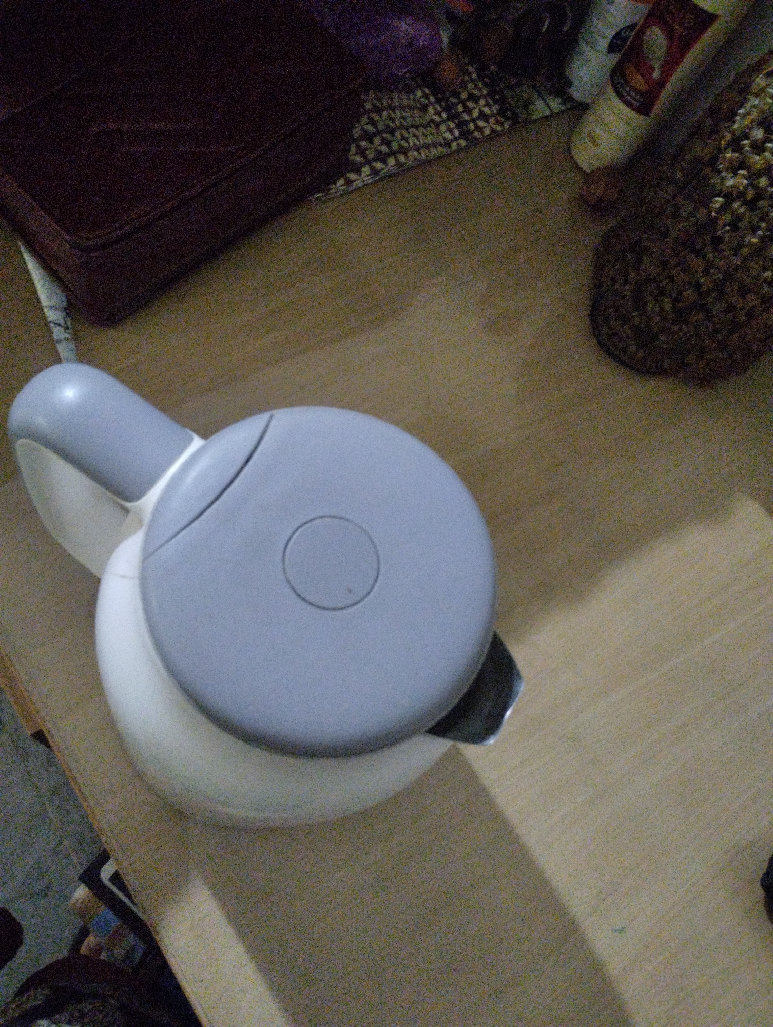 A white electric kettle with a light gray lid and handle is situated on a light-colored wooden surface. The kettle's lid features a circular indentation and a curved line. To the left of the kettle is a dark red upholstered object, possibly a cushion or furniture. In the background, various textured items are visible, including a patterned textile and what appears to be a knitted object. The scene is illuminated by ambient light, casting shadows on the wooden surface. The location is not specified beyond "Unknown, Unknown."