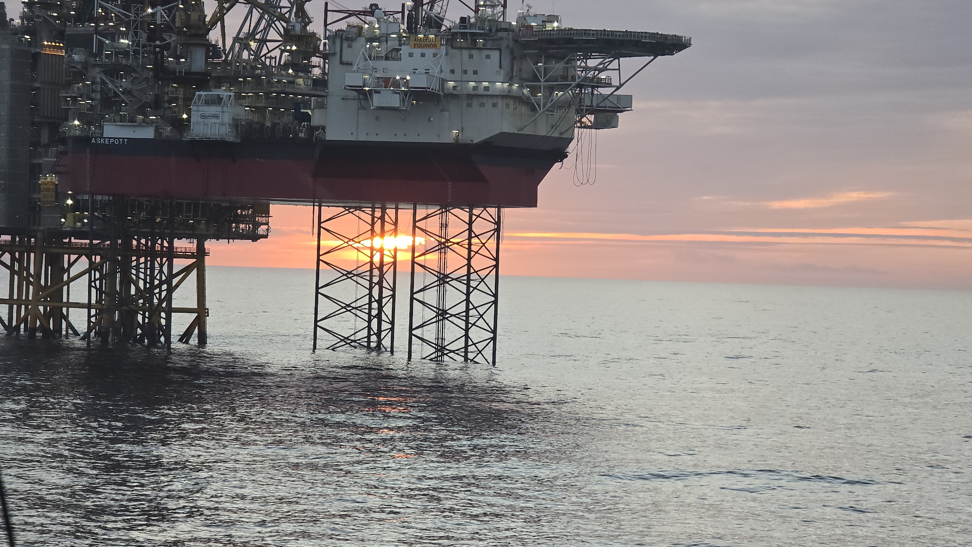 The image depicts the "Askelott" oil platform located in the North Sea, off the coast of Randaberg, Norway. The platform is a large structure with a red and white exterior, supported by a network of steel legs submerged in the water. The sun is setting behind the platform, creating a warm glow in the sky and reflecting off the water's surface. The sea is calm with small ripples. The sky is a mix of pastel colors, primarily pink and light blue. The platform has a helipad on top. The name "ASKEPOTT" is visible on the side of the platform. There are no people visible in the image.