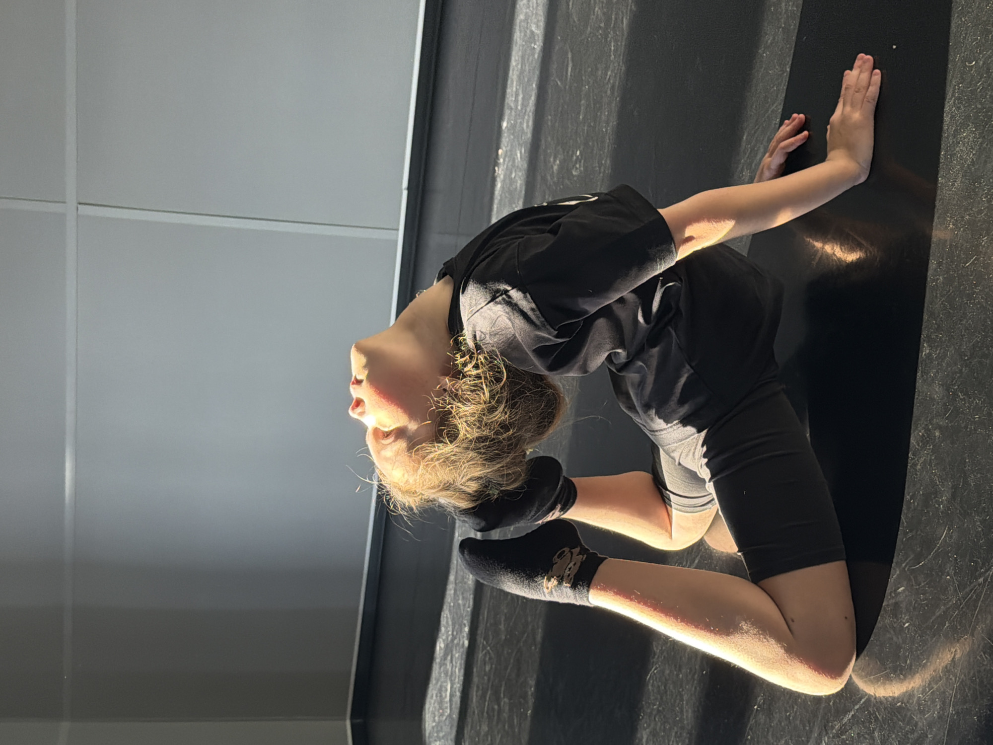 A young person is performing a backbend on a dark floor. The person has on black socks, black shorts, and a black t-shirt. The person's hands are pressed against the floor. The background features a grey wall. The scene takes place in a room with natural light. There is no indication of the location being in Saint Petersburg, Russia.