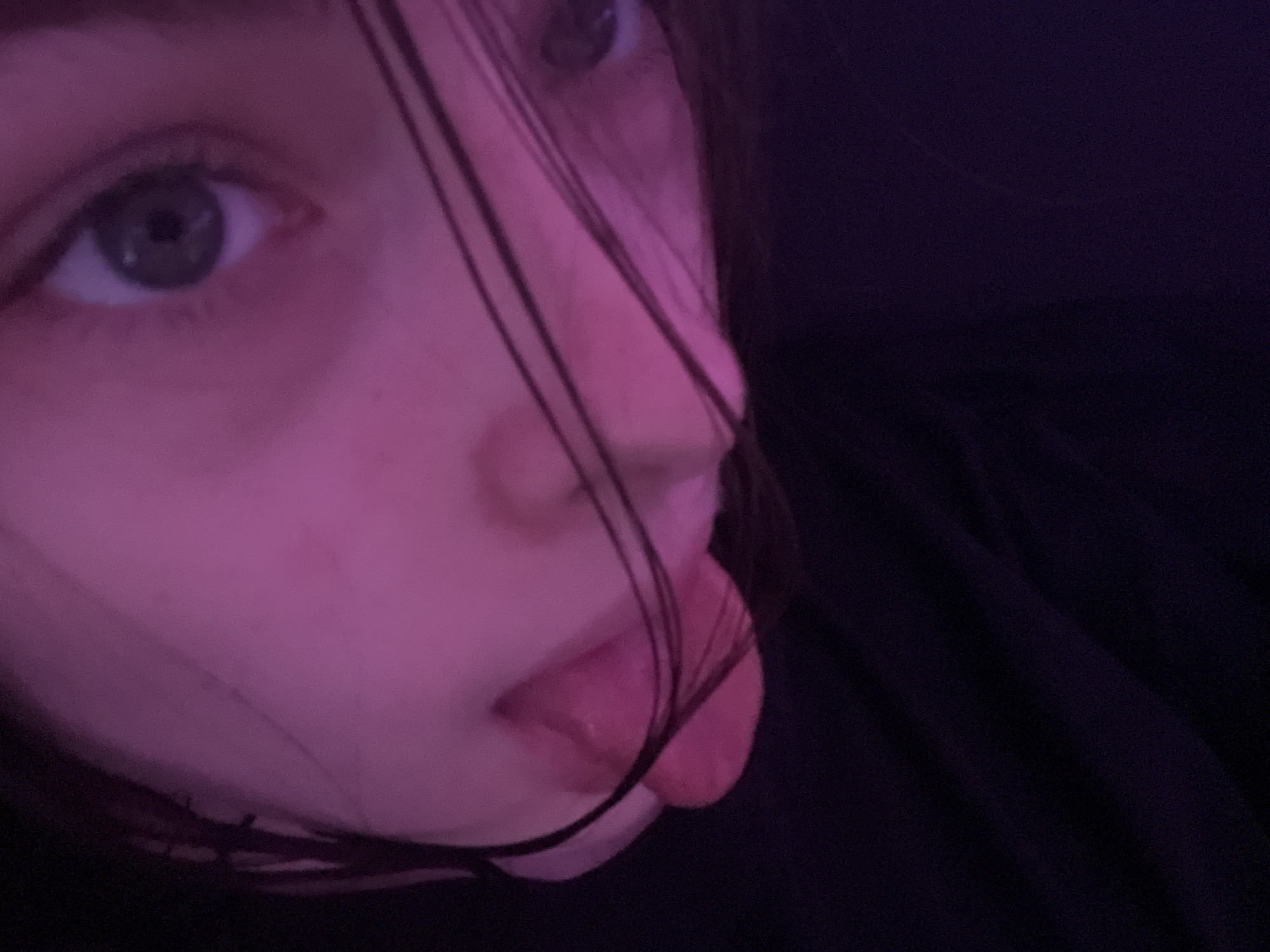 A close-up, low-angle shot depicts a person's face under pink lighting. The individual has fair skin, grey eyes, and dark hair that partially obscures their face. The person is sticking their tongue out. The background is a dark, blurry expanse, likely fabric. No information suggests this photo was taken in Saint Petersburg, Russia.