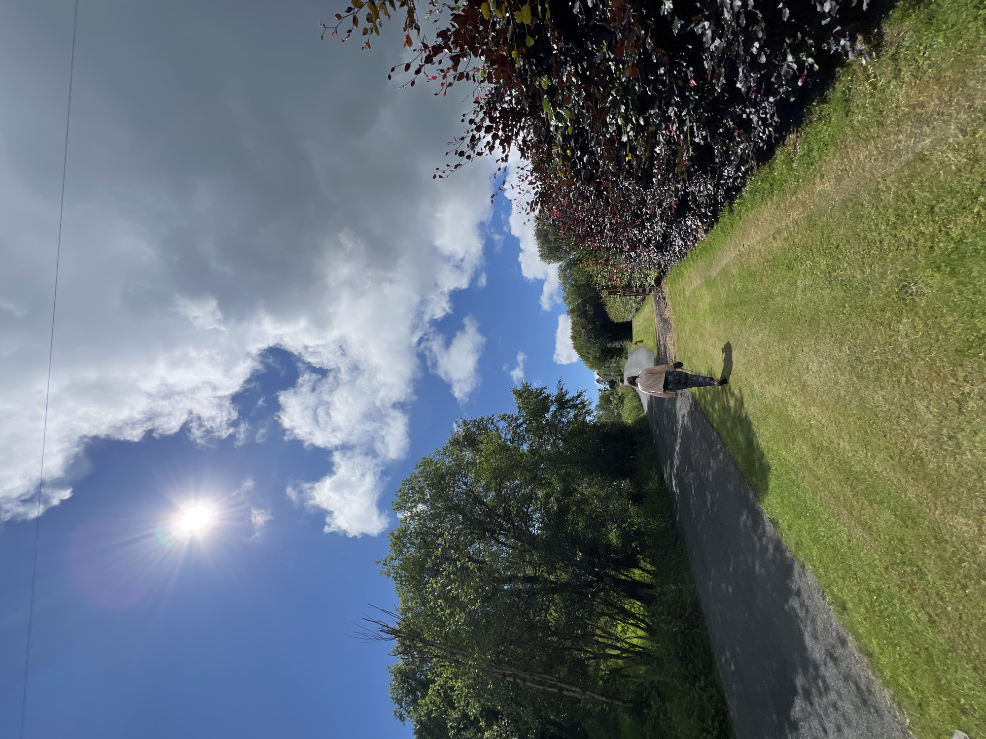 The image depicts a sunny day with scattered clouds in a rural setting. A person is walking on a paved path bordered by a grassy area and dense foliage. The sun is bright, casting shadows and creating a lens flare. The scene suggests a leisurely walk outdoors, possibly in a park or along a country lane. The location context of Mountcharles, Ireland, implies a temperate climate and a landscape with lush greenery. There is no visible text in the image. The overall mood is serene and pleasant, indicative of a good weather day.