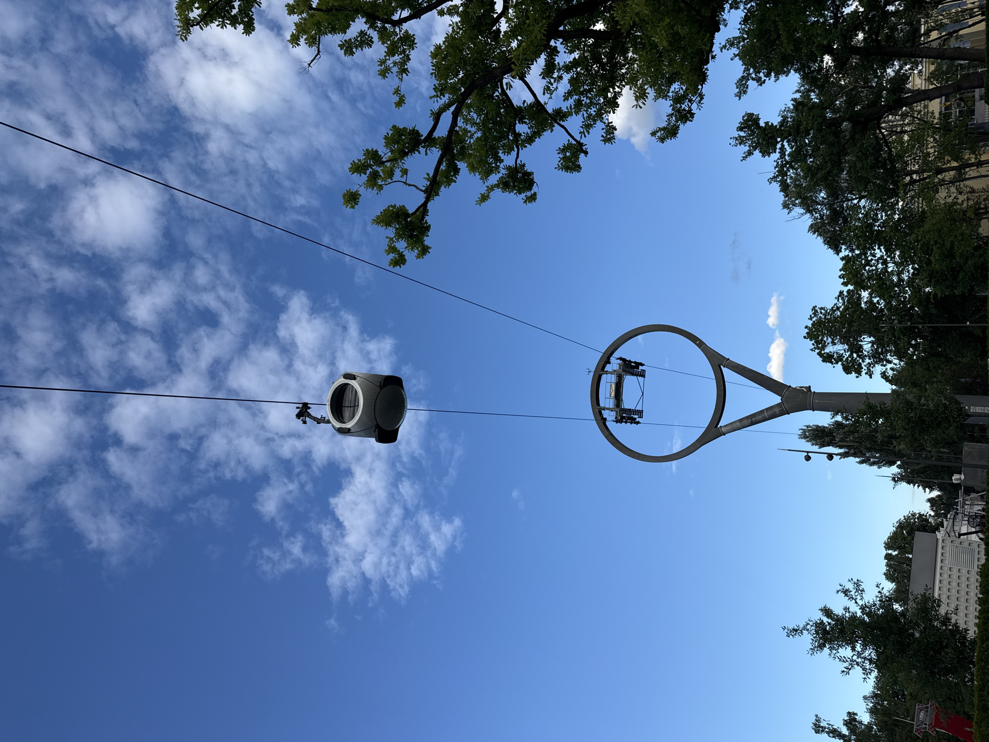 The image depicts a clear blue sky with scattered white clouds, a common sight over Moscow, Russia. In the foreground, a cable car hangs suspended from a wire, its metallic exterior reflecting the sunlight. Below it, a circular structure, part of the cable car's support system, is visible. Trees with lush green foliage frame the scene, suggesting a park or outdoor recreational area. The overall impression is one of a sunny day in an urban setting, likely during the warmer months.