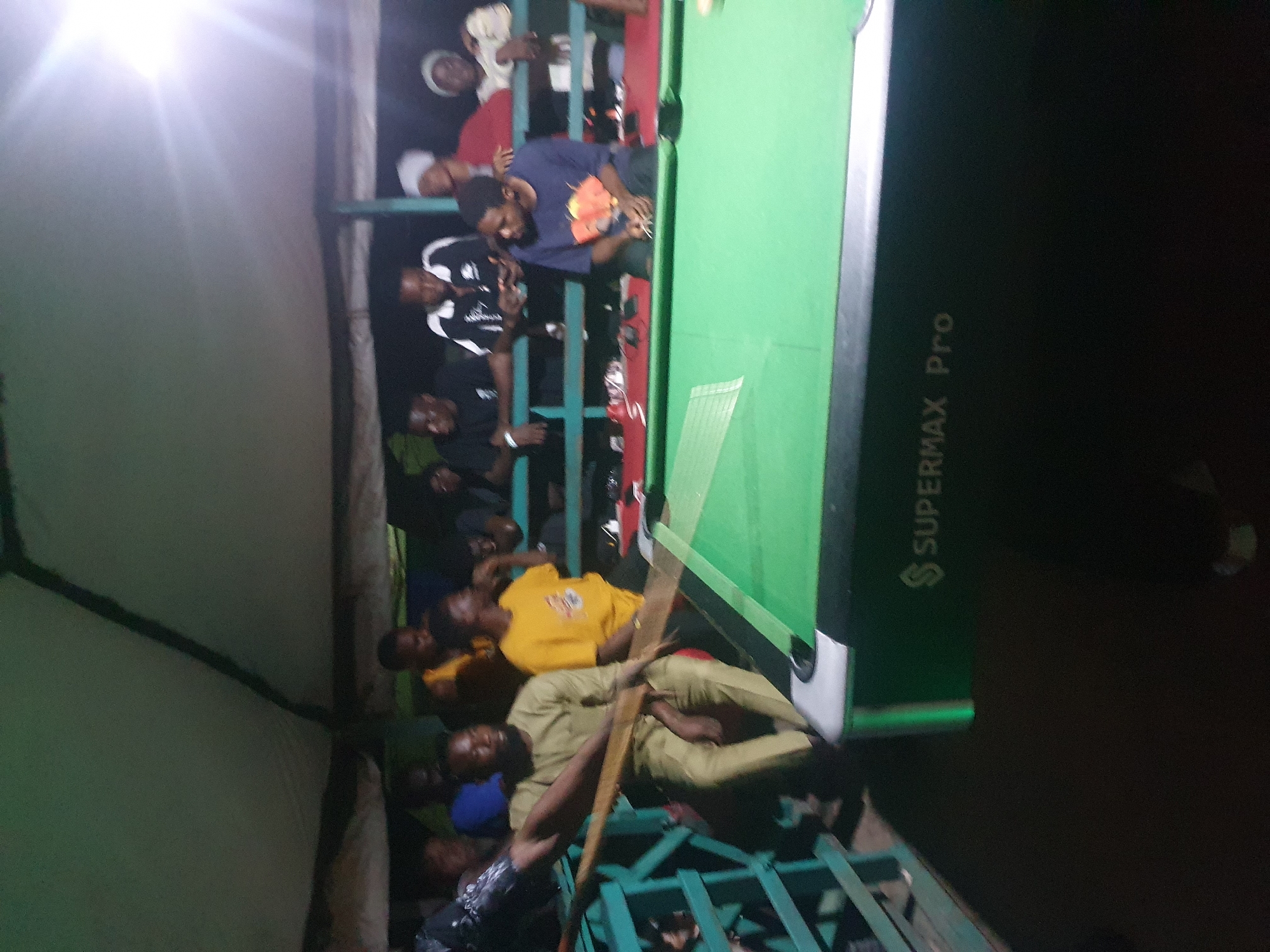 The image depicts a group of young men gathered around a pool table, likely playing a game of billiards. The scene is dimly lit, suggesting it is nighttime. The men are dressed casually, with some wearing t-shirts and others in sportswear. They appear to be engaged and focused on the game, with some observing and others actively participating.
The pool table itself is green, and one of its sides has text that reads "SUPERMAX Pro". The men are positioned around the table, some standing and others leaning over to take shots. The overall atmosphere seems relaxed and social, with a sense of camaraderie among the individuals.
The environment appears to be an outdoor or semi-outdoor setting, possibly a recreational area or a local spot in Lokoja, Nigeria, where such games are common. The background is dark and out of focus, providing little detail about the surroundings. The lighting suggests artificial light sources, common for nighttime activities. No specific emotions are clearly discernible due to the image quality and lighting, but the focused posture of some individuals suggests concentration on the game.