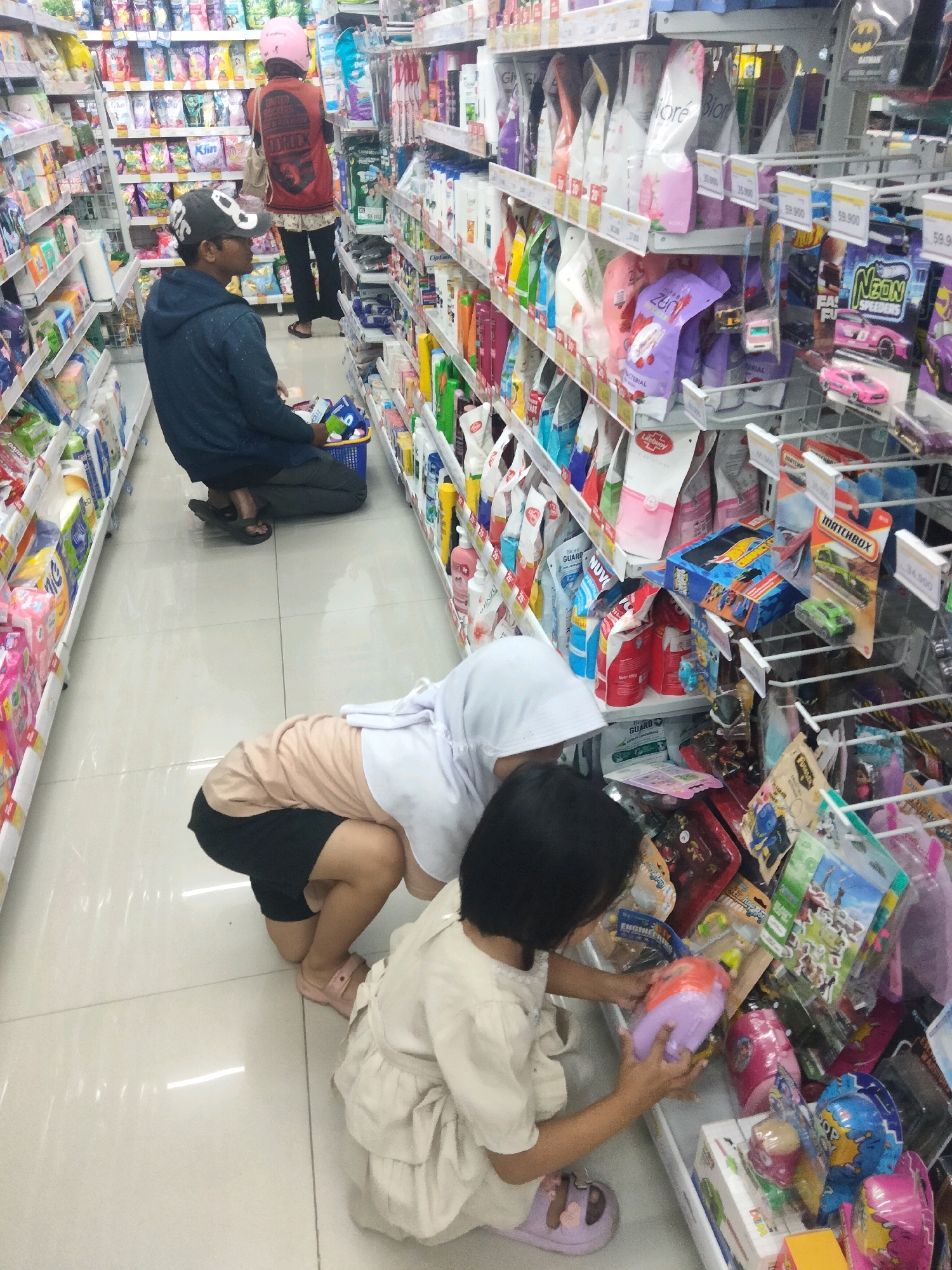 In a brightly lit aisle of a convenience store in Suka Sari, Indonesia, two children and one adult are engaged in shopping. The adult, wearing a dark blue hoodie and a baseball cap, is crouching and looking down at a blue shopping basket filled with items. To their right, a child in a light beige dress and a white hijab is kneeling and reaching for a toy. Another child, partially visible, is also kneeling and wearing a light pink shirt and dark shorts. The shelves surrounding them are stocked with a variety of products, including packaged goods, toiletries, and toys. The floor is tiled and reflective.