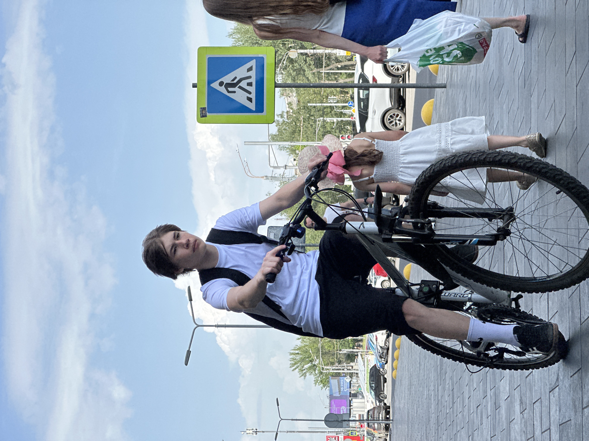 The image shows a young person riding a bicycle on a city sidewalk. The rider is wearing a white t-shirt, black shorts, white socks, and black sneakers. They also have a black backpack. A crosswalk sign is visible in the background. Other people are walking on the sidewalk nearby. In the distance, cars and buildings can be seen, characteristic of an urban area. Based on visual cues like the crosswalk sign design, architectural styles, and local license plates on cars, this image was taken in the city of Nizhny Novgorod, Russia.