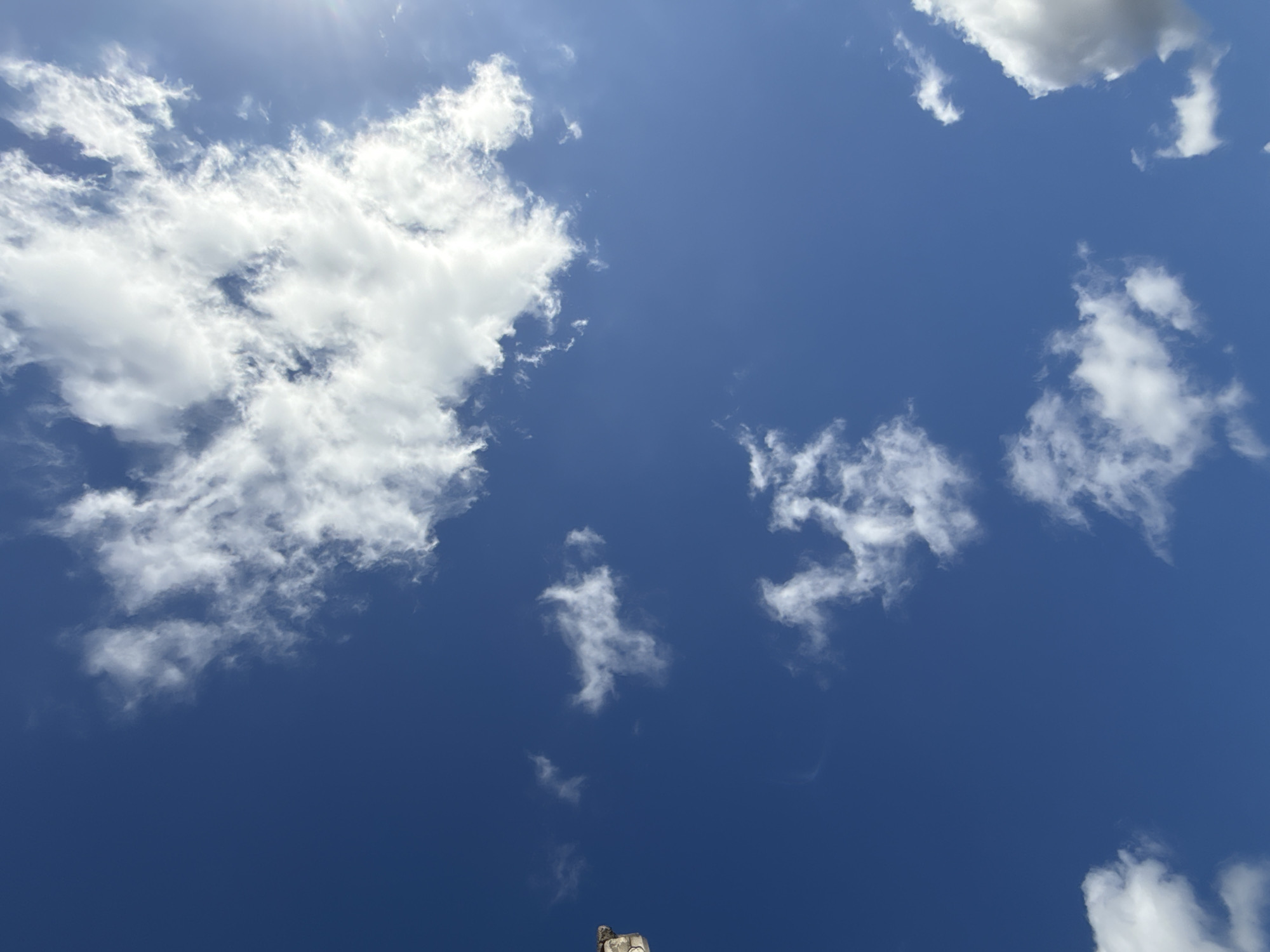 The image shows a bright blue sky with scattered white clouds. The sun is visible in the upper right corner, creating a lens flare. A small portion of a stone structure is visible in the upper left corner of the image. The scene appears to be an outdoor environment, possibly during the day. No people or specific actions are depicted in the image. The location in Walkenried, Germany is not evident from the visual information provided in the image.