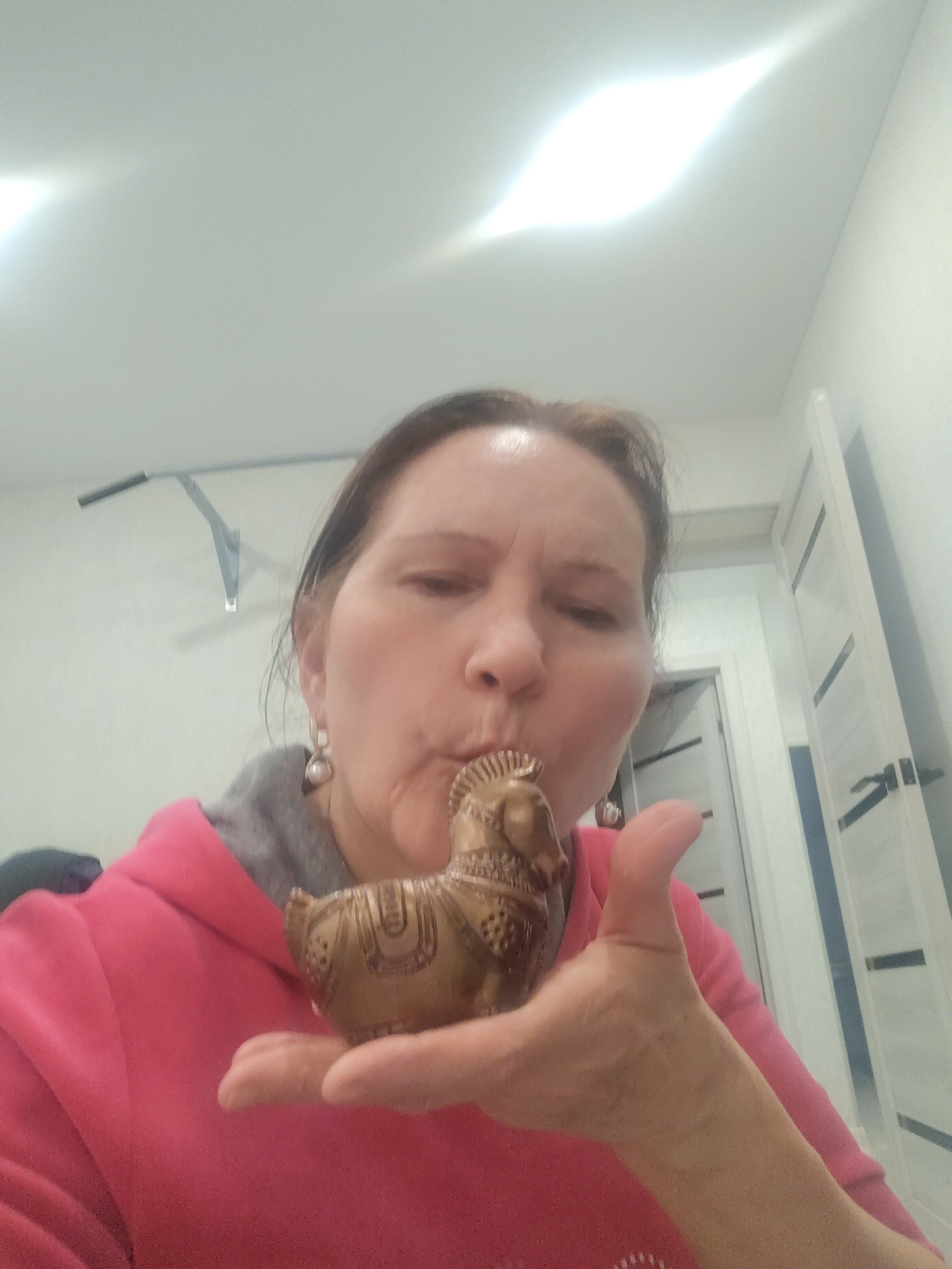 A middle-aged woman wearing a pink hoodie and pearl earrings is indoors, possibly in a residence. She is holding a small, detailed, bronze-colored horse figurine in her left hand, which is positioned close to her face as if she is about to kiss it or blow on it. The background shows a white wall, a door with dark accents, and a pull-up bar attached to the wall. According to the location information, this scene may be located in the city of Sarapul, Russia.
