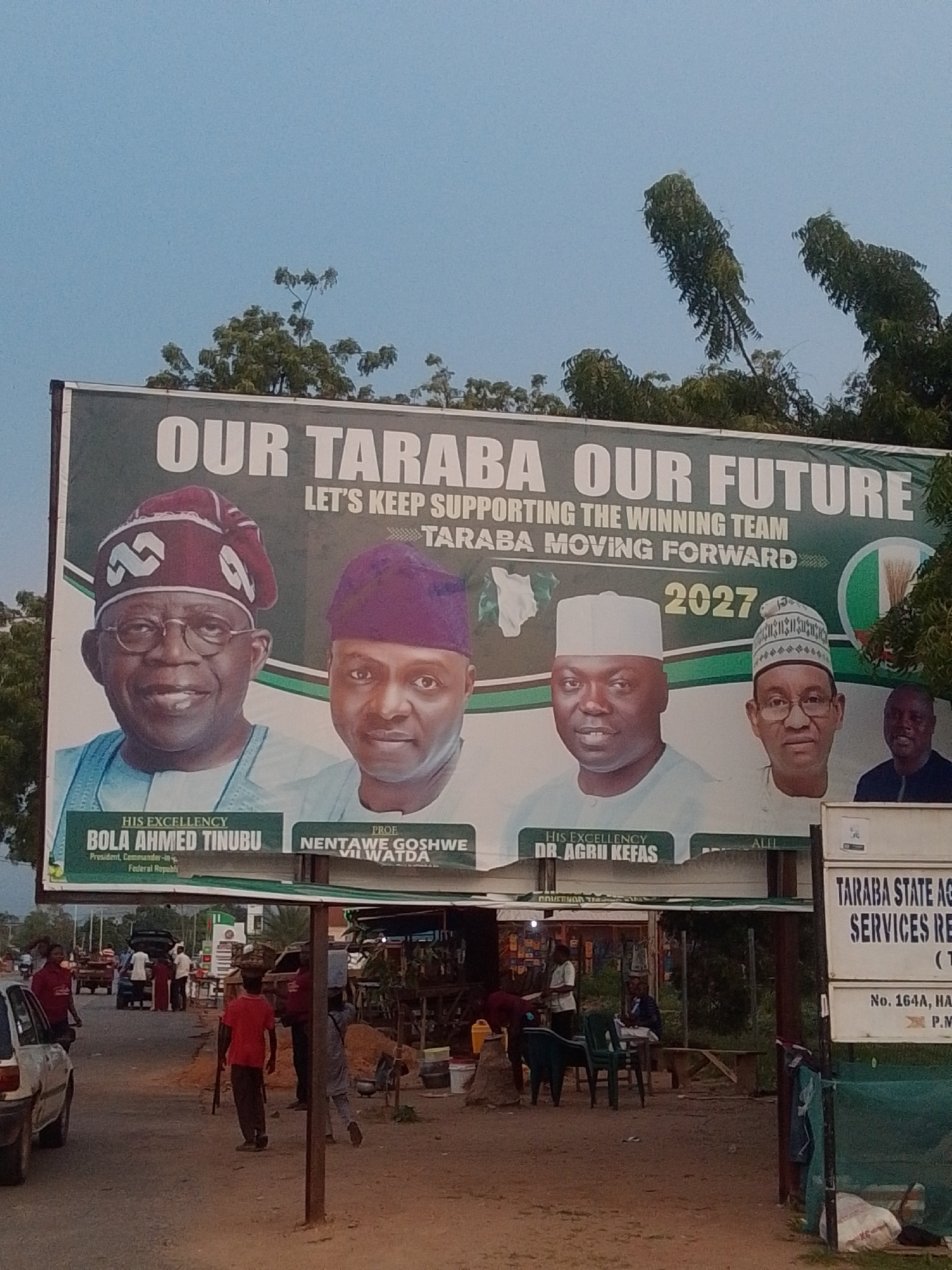 The image shows a billboard with the text "OUR TARABA OUR FUTURE LET'S KEEP SUPPORTING THE WINNING TEAM TARABA MOVING FORWARD 2027". The billboard features portraits of four men: Bola Ahmed Tinubu, Nentaw Goshwe VII Watda, Dr. Agbu Kefas, and Ali. Below their portraits, their titles are listed. The scene appears to be in Jalingo, Nigeria, indicated by the presence of a sign for "TARABA STATE AGENCY SERVICES RE" which is likely "Taraba State Agency for Agricultural Services" or a similar government entity. The billboard is mounted above a roadside stall or small market area, with people and vehicles visible in the background. The lighting suggests it is daytime, possibly late afternoon.