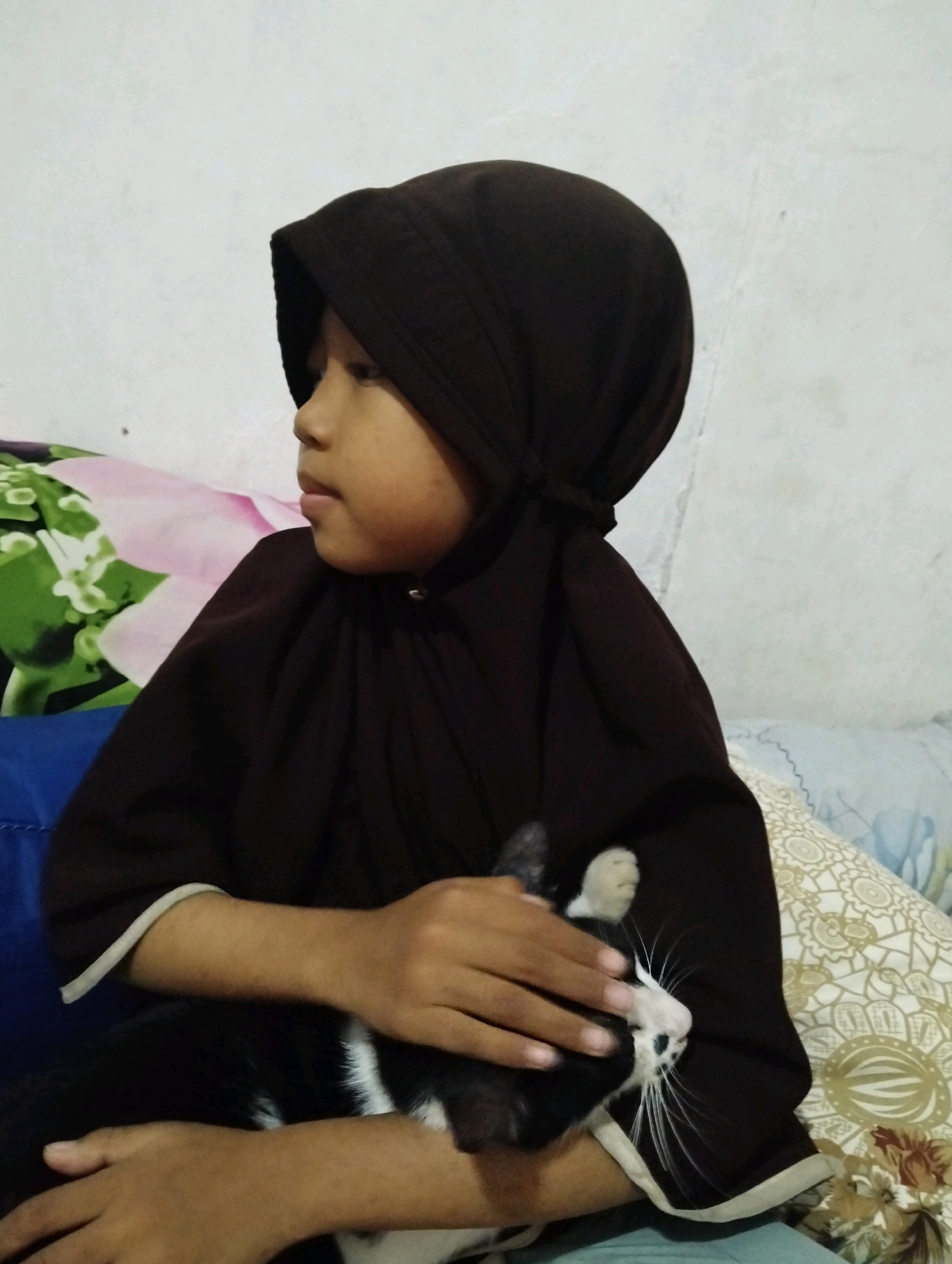 A young person wearing a dark brown hijab and clothing sits with a black and white cat. The person's hands cradle the cat. The cat's head rests on the person's arm, and its whiskers are prominently displayed. The background consists of a white wall and pillows with floral patterns. The scene is indoors, likely in a residential setting in Cileungsi, Indonesia.