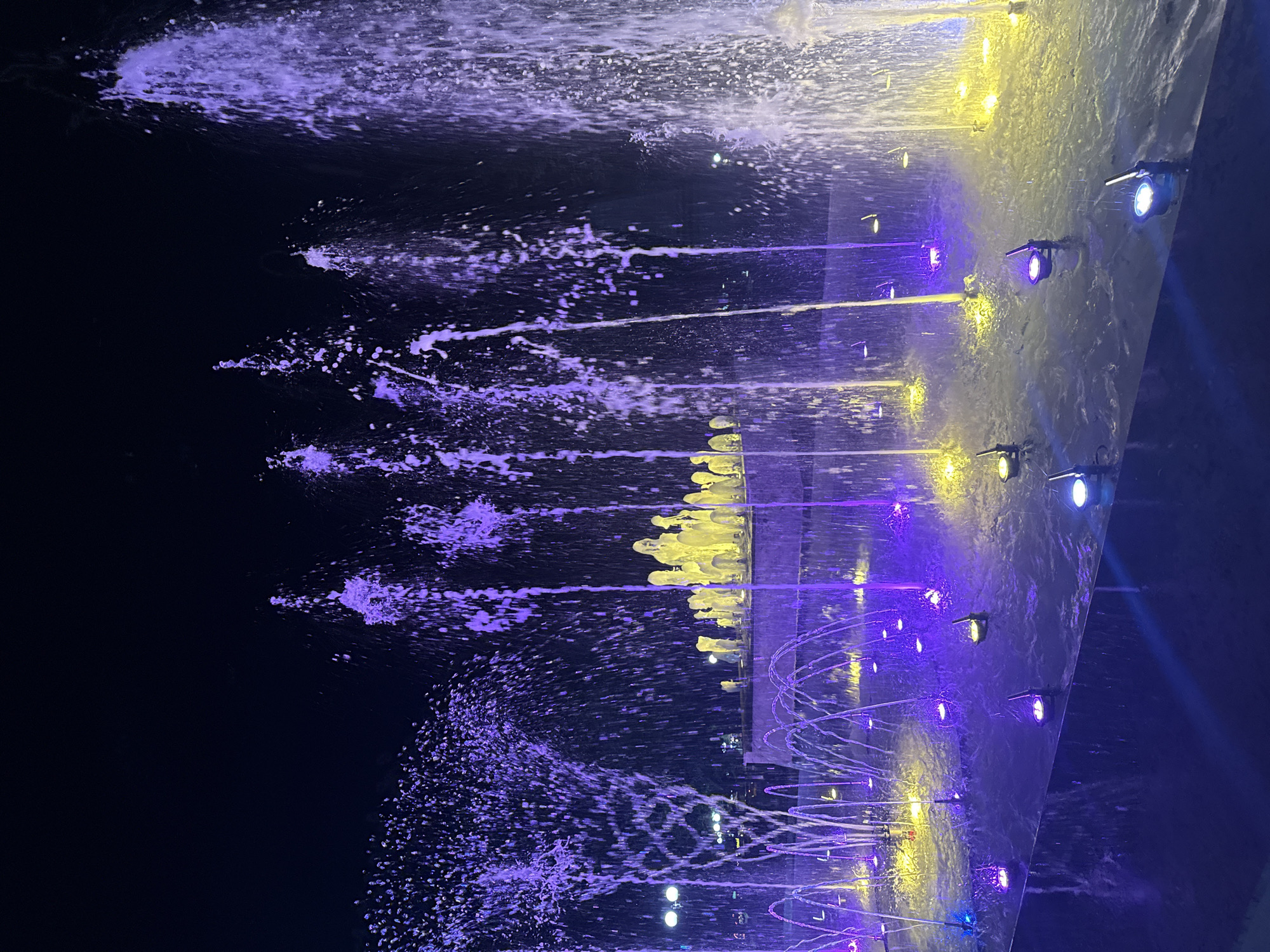 A fountain is illuminated by purple and yellow lights at night. The fountain has many jets of water spraying upwards. In the background, there are more water jets and some yellow lights. The area around the fountain is dark, suggesting it is nighttime. The scene is likely in Stara Zagora, Bulgaria, given the context of the query. There are no people visible in the image. The fountain appears to be in an outdoor public space.