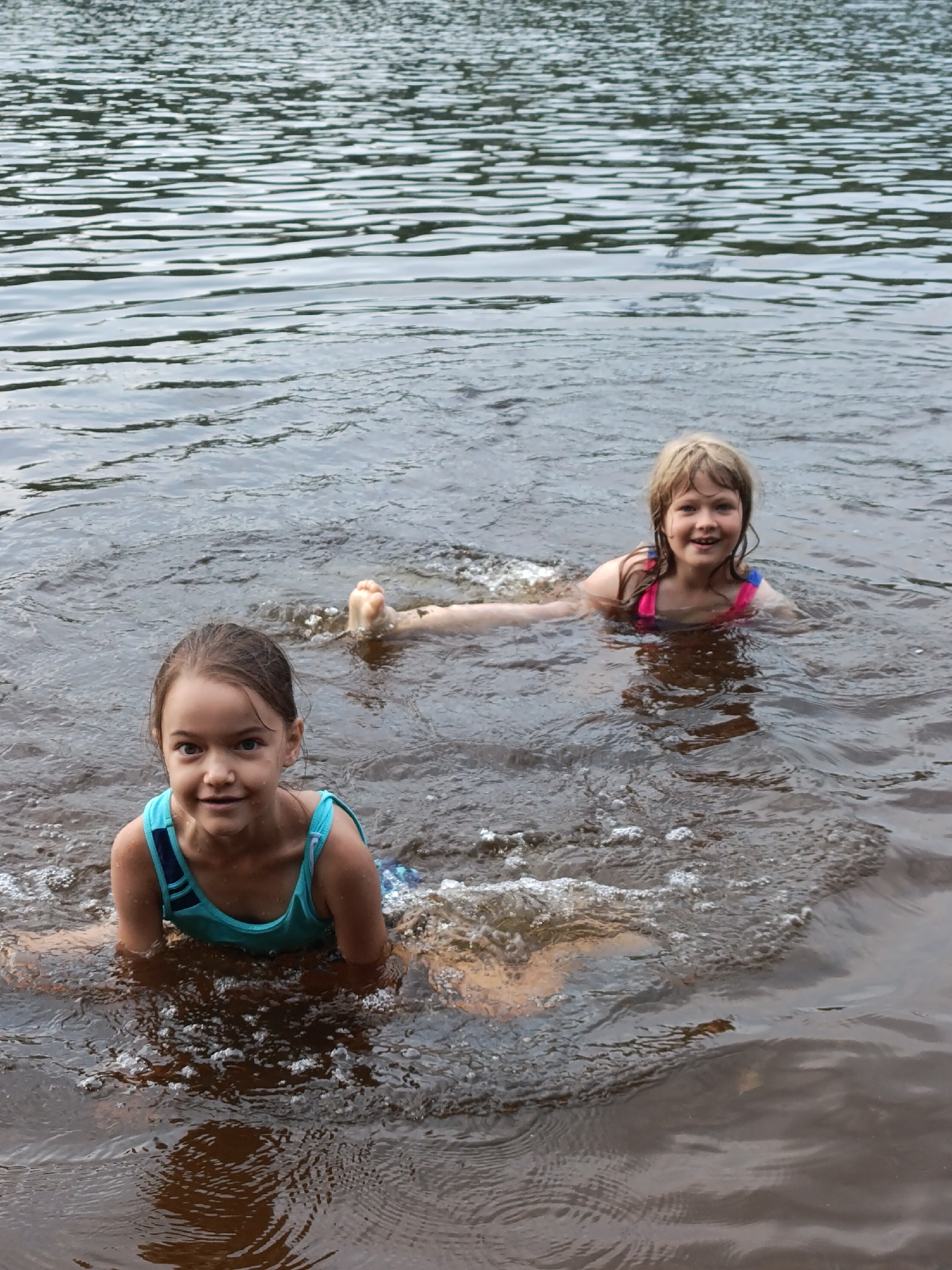 Two young girls are in the water, seemingly enjoying themselves. The girl in the foreground, on the left, is closer to the camera and is wearing a blue and black swimsuit. She is looking directly at the camera with a smile. Her hair is dark and pulled back. The girl in the background, on the right, is also in the water and appears to be swimming or playing. She is wearing a pink and blue swimsuit and has lighter colored hair that is wet and spread out around her. Her face is turned towards the camera, and she has a joyful expression. Her right leg is visible above the water. The water itself appears somewhat murky and has ripples and some small waves, suggesting movement. Bubbles are visible around the girls. The overall setting is an outdoor body of water, likely a lake, river, or pond, given the calm appearance of the water and the natural lighting. The time of day appears to be daytime, judging by the light. There is no visible text in the image.