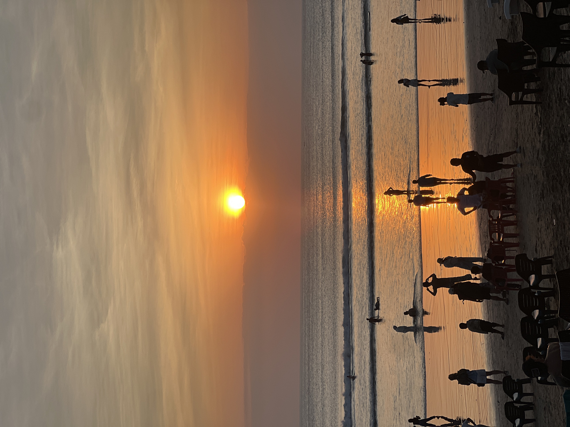 This image depicts a sunset scene at Kuta Beach in Bali, Indonesia. Silhouetted figures of people are visible on the wet sand, some wading in the shallow water, others standing and observing the ocean, and a few are seated on beach chairs in the foreground. The sun, a bright orb, is low in the sky, casting a warm orange glow across the water and the clouds. Gentle waves are rolling onto the shore, and their reflections shimmer on the wet sand. The scene captures the tranquil atmosphere of a beach at dusk.