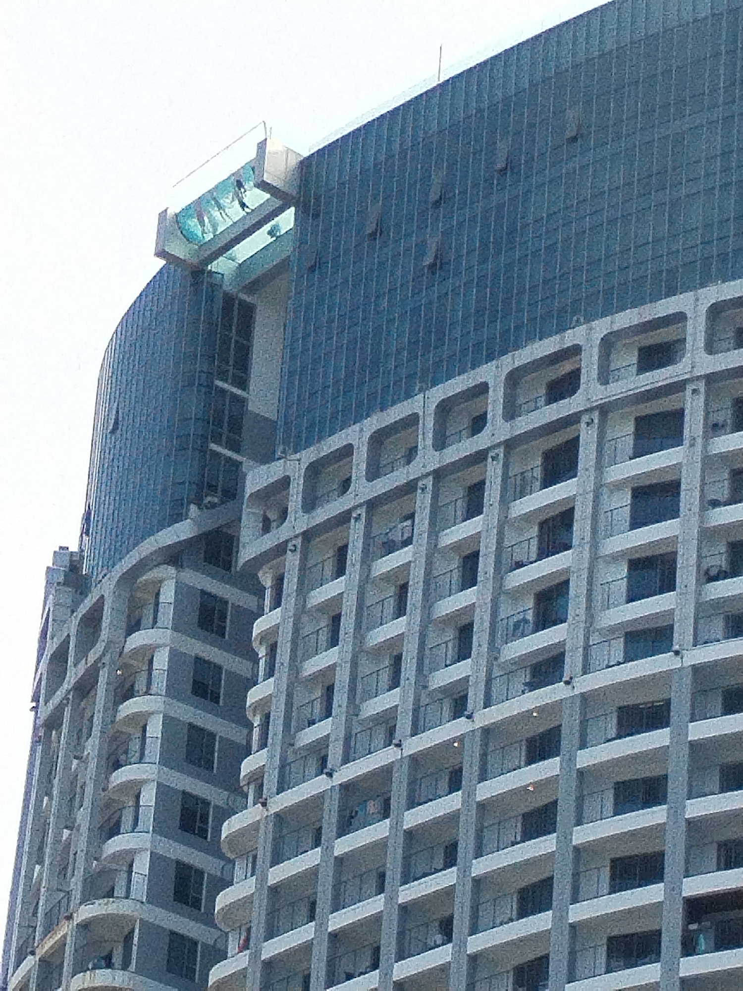The image shows a close-up of a modern skyscraper in Nha Trang, Vietnam. The building is characterized by its curved, glass facade and multiple levels of balconies. A striking feature is a glass-enclosed swimming pool cantilevered from the top of the building, appearing to be suspended in mid-air. The sky is overcast and white, suggesting it is daytime. The lower portion of the building exhibits a repetitive pattern of balconies and windows.