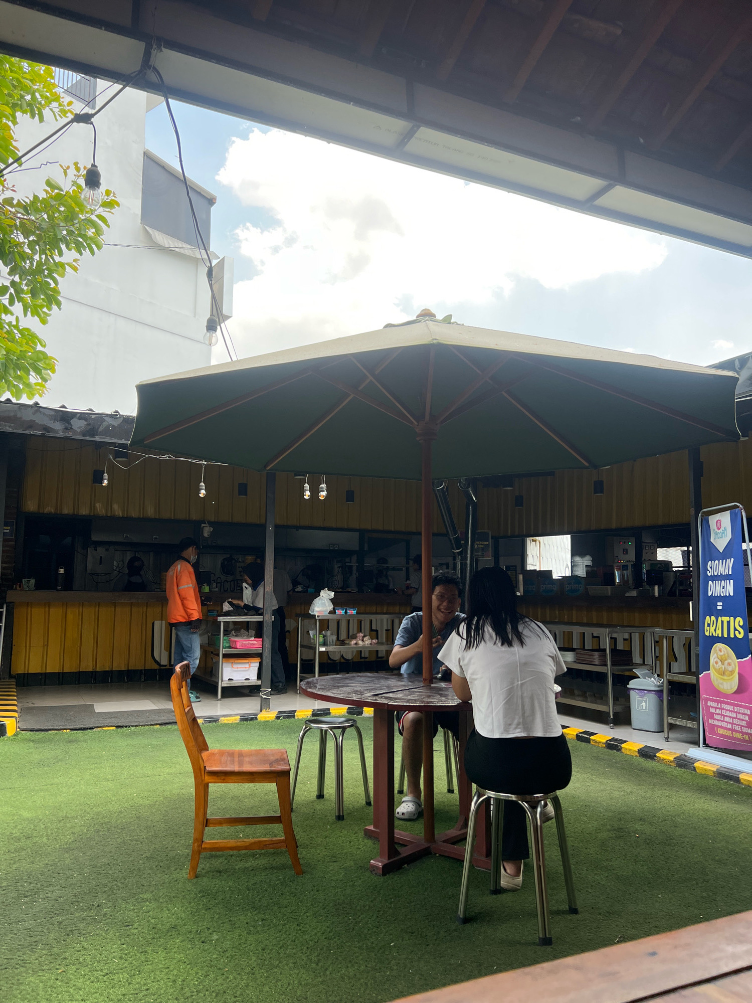 The image depicts an outdoor seating area of a food establishment in Yogyakarta, Indonesia, possibly a "warung" or food stall, characterized by its casual and open-air setup. In the foreground, a wooden chair sits on artificial green turf, facing a round wooden table with two metal stools. Two individuals are seated at the table, facing away from the camera. One is a woman with long dark hair wearing a white top and black pants. The other, a man, is partially visible, wearing a blue t-shirt and shorts, smiling and looking towards the woman.
The seating area is shaded by a large, olive-green umbrella. Behind the umbrella, a yellow-walled building with an open-fronted kitchen area is visible. Several individuals can be seen working behind the counter. A sign with Indonesian text and an image of food is prominently displayed to the right of the umbrella.
The environment suggests a warm climate, with natural light filtering through a partly cloudy sky. String lights are suspended from the overhead structure, suggesting it might be used in the evening as well. The overall impression is of a relaxed, informal dining space, typical of many eateries in Yogyakarta.