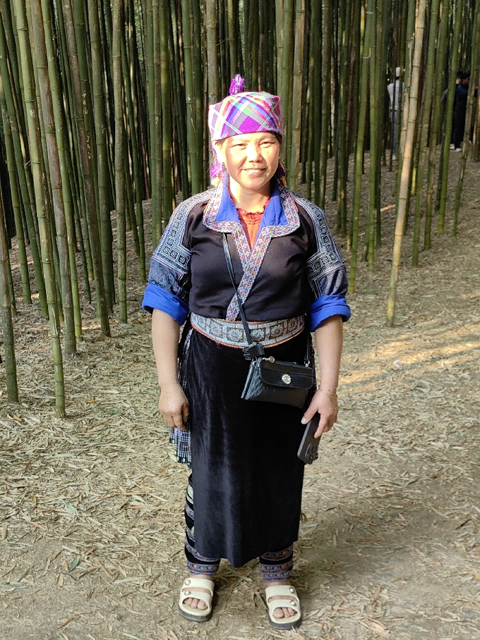 An adult female, estimated to be in her 40s to 50s, stands centered within a dense bamboo forest in Mù Cang Chải, Vietnam. She faces the camera directly, displaying a slight smile. She wears traditional Hmong attire: a plaid headwrap in shades of pink, purple, and green with a purple tassel; a black long-sleeved shirt featuring blue and white patterned embroidery on the shoulders and cuffs, with a blue collar and red inner garment visible at the neckline; a wide, silver-and-black patterned belt with red trim; a black velvet-like long skirt layered over patterned leggings; and light-colored open-toed sandals with two studs on each strap. A small black crossbody bag with a circular silver clasp hangs at her right hip, and a dark-colored smartphone is held in her left hand. The ground is covered with dried bamboo leaves and debris. In the distant background, partially obscured by the bamboo stalks, are two indistinct figures.