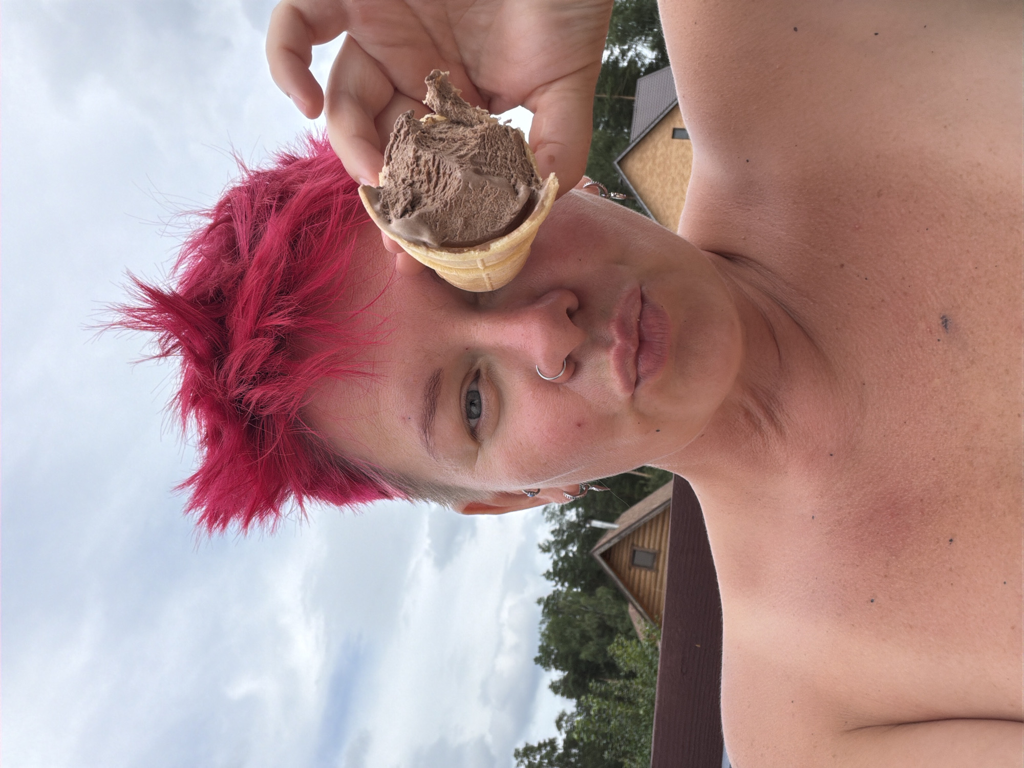 In an outdoor selfie, a person with vibrant pink, spiky hair holds a chocolate ice cream cone to their right eye, obscuring it. They have a nose ring and ear piercings. The background shows a cloudy sky, trees, and the rooftops of wooden houses, consistent with a suburban or rural setting. The individual is shirtless, with their shoulders and upper chest visible, displaying some freckles and what appears to be a tan line or redness on their chest. The context suggests a casual moment outdoors, possibly in Chaikovsky, Russia, given the architectural style of the houses and vegetation which are characteristic of the region.