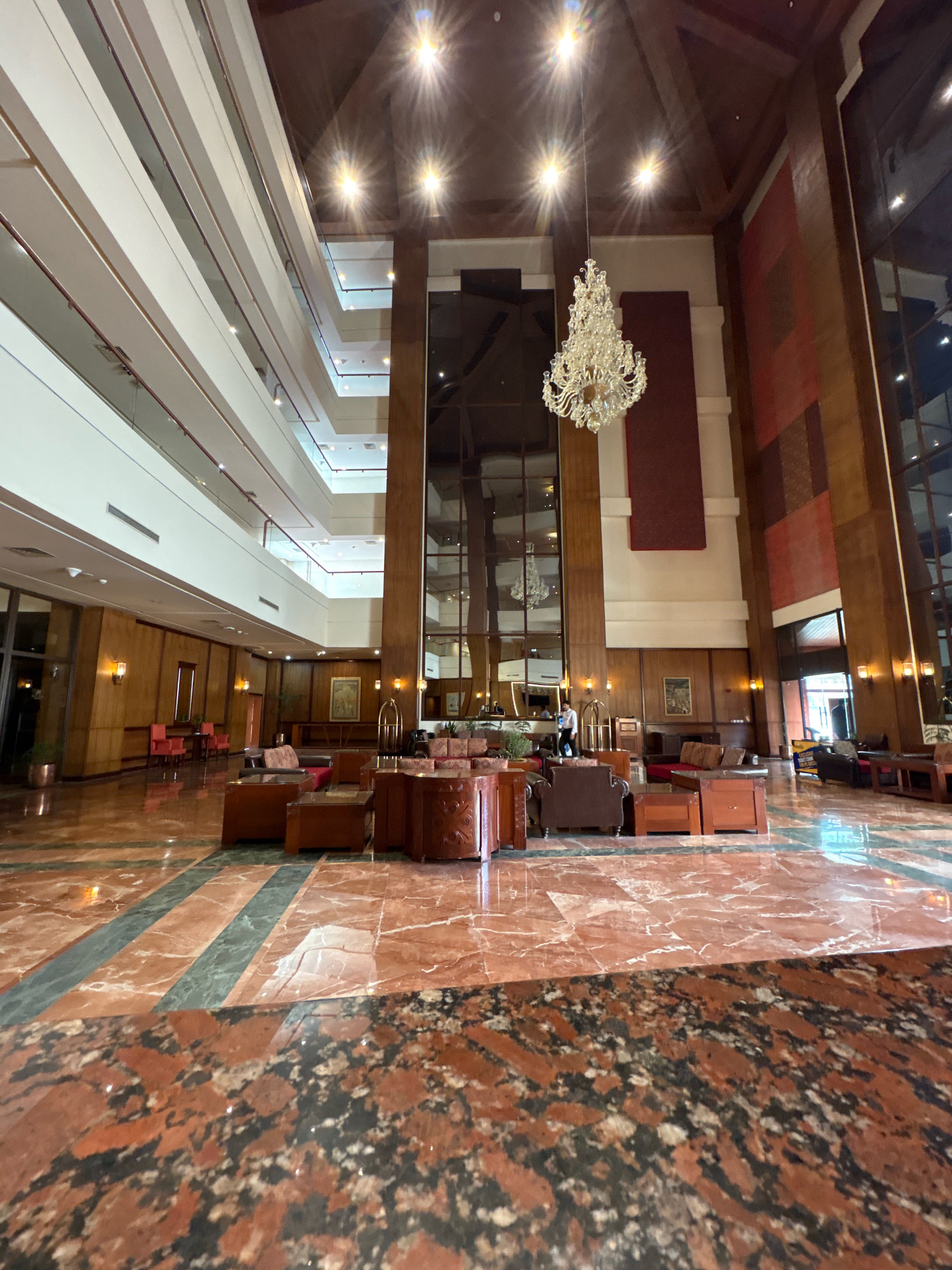 The image shows the interior of a grand hotel lobby, possibly in the city of Malam Jabba, Pakistan, though this cannot be confirmed from the visual information alone. The lobby features a multi-story atrium with white balconies and wooden paneling on the walls. A large, ornate crystal chandelier hangs from the ceiling, illuminated by bright lights. The floor is a polished marble with a complex pattern of red, green, and black stones. In the center of the lobby, a seating area with brown leather sofas and wooden tables is visible. To the right, a hotel employee in a white shirt is attending to a luggage cart. Several decorative paintings hang on the wooden walls. The overall impression is one of opulence and spaciousness.