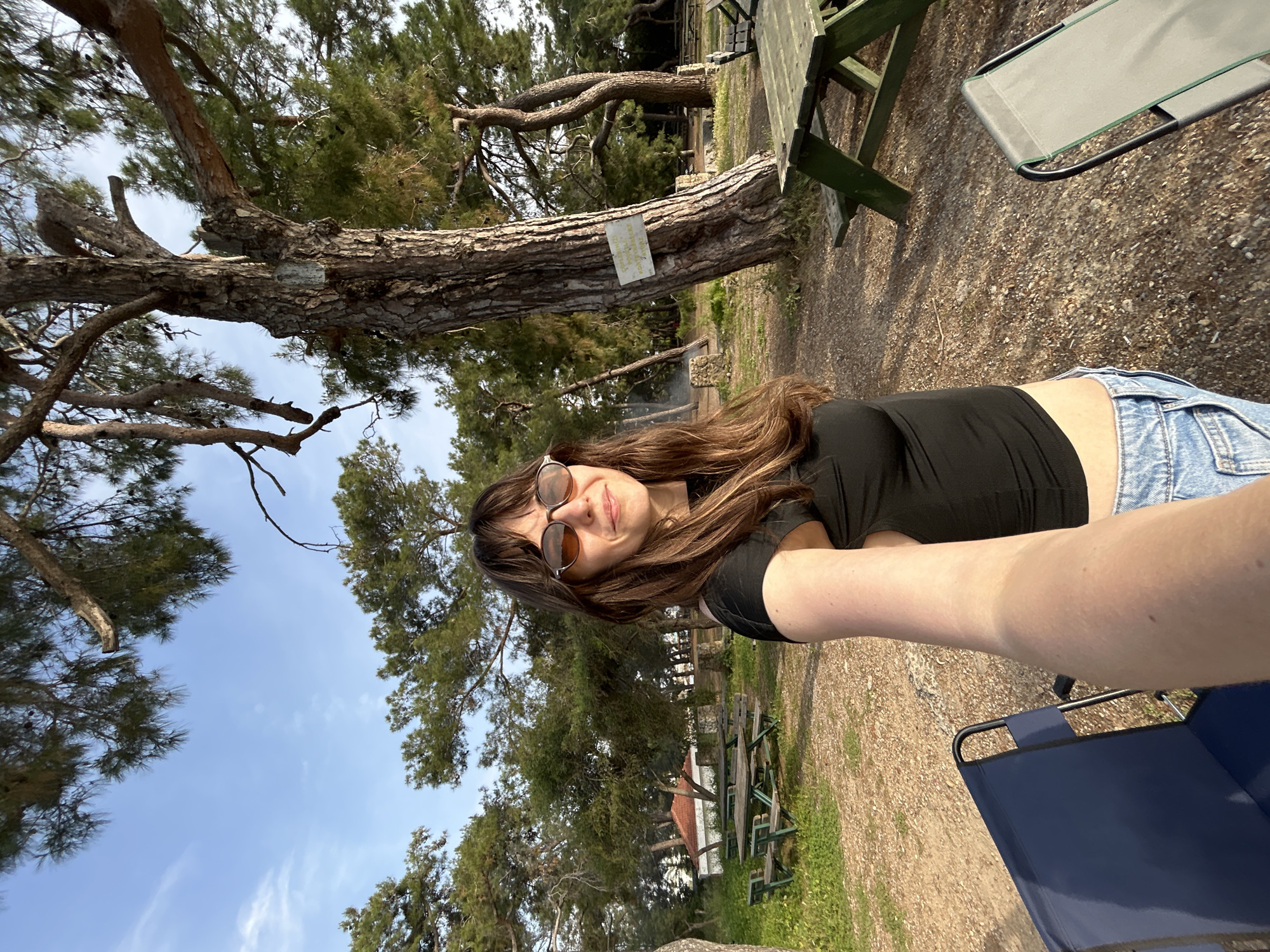 A woman in sunglasses, a black top, and blue denim shorts takes a selfie at a park, possibly in Alanya, Turkey. The scene is outdoors, with trees and a picnic table in the background. The sky is partially visible, with some clouds. The ground is covered with dry leaves and gravel. A blue chair is partially visible in the foreground. The photo appears to be taken at an angle. There is a white sign on a tree trunk.