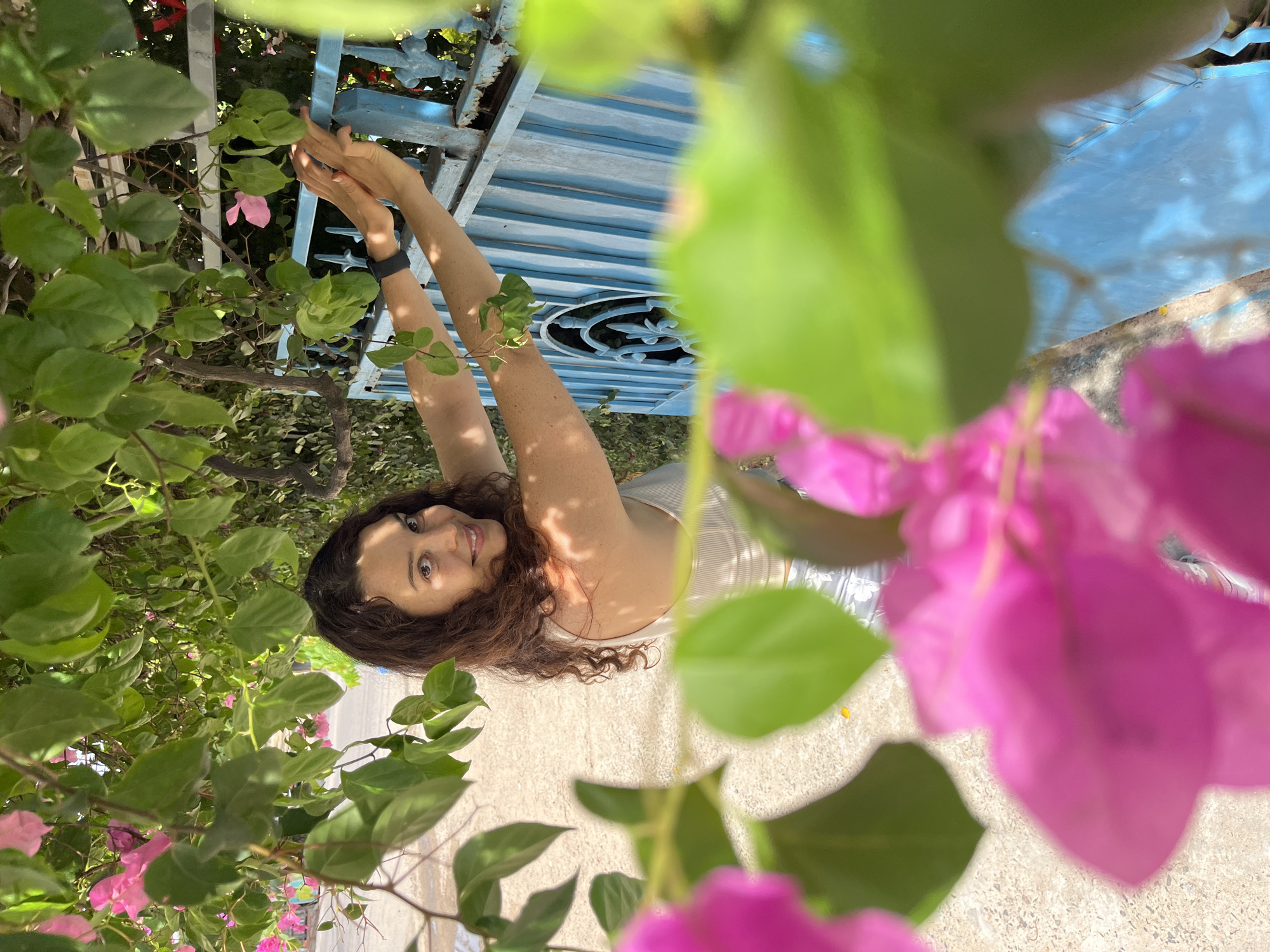 A woman with dark curly hair smiles at the camera, positioned under the dappled shade of green leaves and pink bougainvillea flowers. She is leaning against a faded blue metal fence, her hands resting on the top bar of the fence. She is wearing a white tank top and patterned shorts. The fence has intricate scrollwork visible through its bars, and appears to be slightly rusted in places. Behind the fence, foliage is visible, suggesting an outdoor setting. The ground is a sandy-colored path. The image is taken in Mũi Né, Vietnam, a coastal resort town known for its beaches and sand dunes.