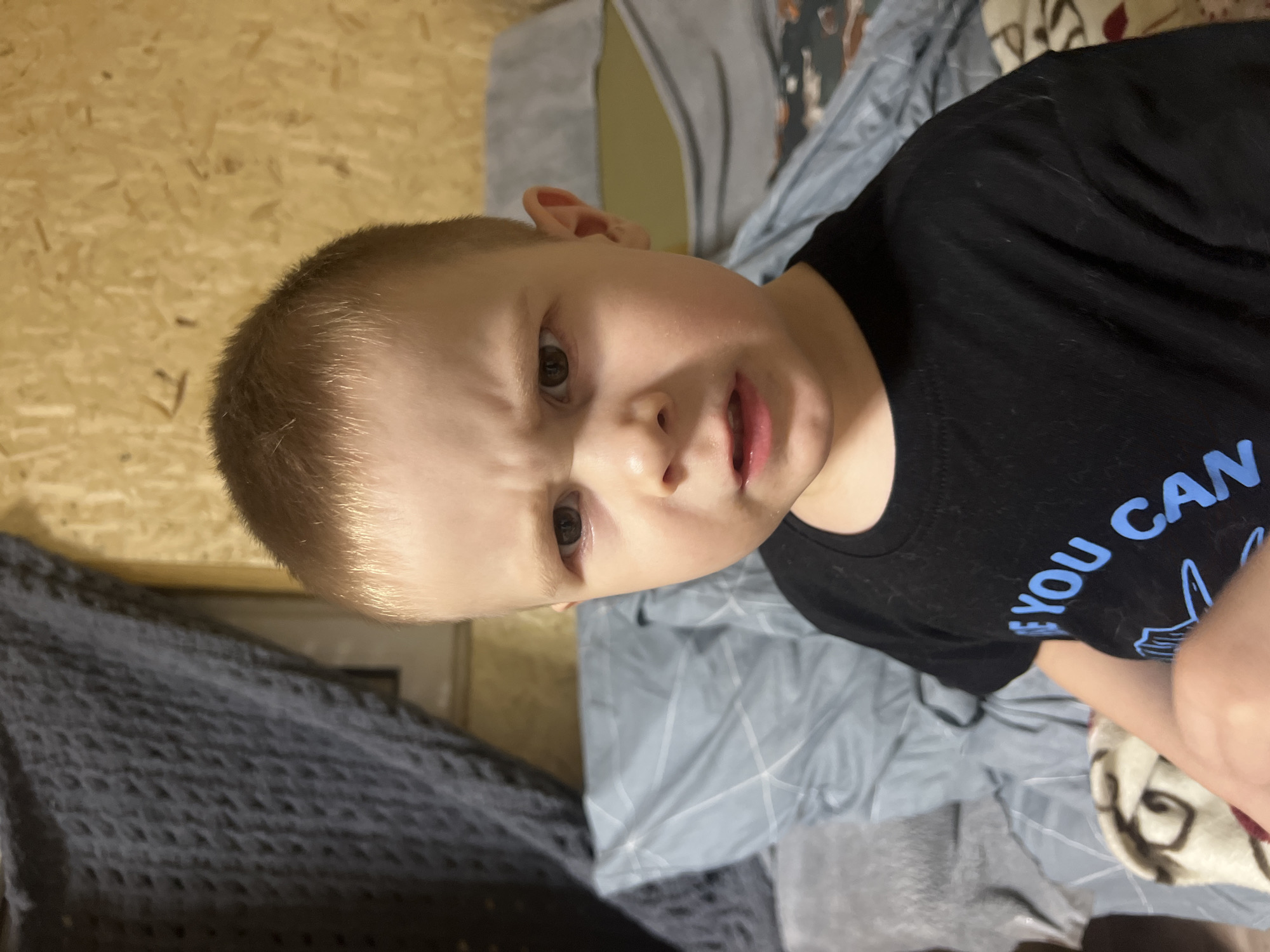 A young boy with blonde hair is positioned in front of a background consisting of a light-colored wooden wall and layered bedding. He is wearing a black t-shirt with blue lettering. The boy's expression appears serious, with furrowed brows. The location is unspecified in the provided information, and cannot be assumed to be Chaikovsky, Russia.