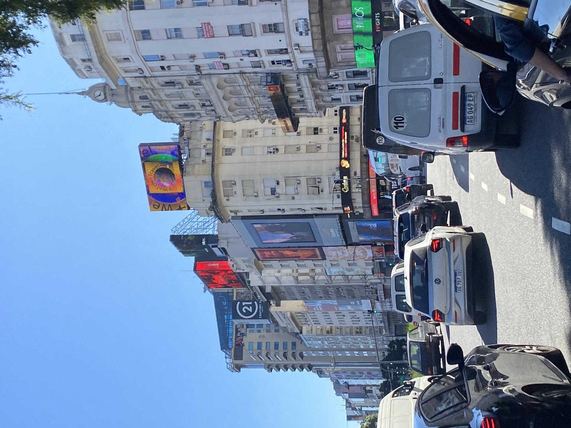 This is a daytime street scene in Buenos Aires, Argentina, showing traffic on a wide avenue lined with tall buildings. The sky is clear blue. The buildings feature prominent digital billboards and advertising signs. In the foreground, there are several vehicles, including a white van with the license plate "AG 049 EP," a white Volkswagen with license plate "AG 787 TU," and other cars. The vehicles appear to be in slow-moving traffic or a standstill. Some signs visible include "CENTURY 21," "KFC," and a digital board with "WeLove" displayed. Other signs are commercial advertisements. A building with a clock tower is seen in the distance, and the general architecture is in a European style. The scene depicts a bustling urban environment.