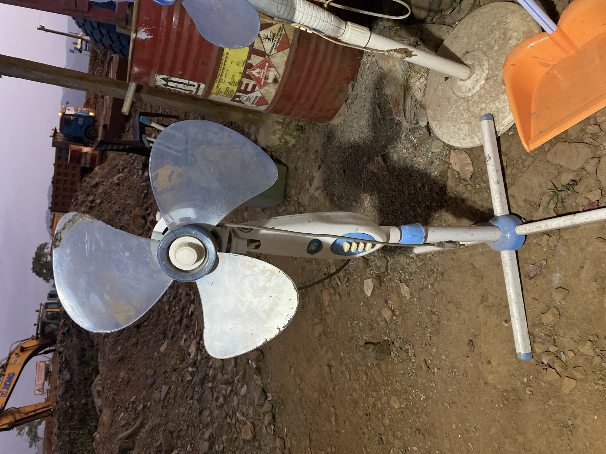 A close-up, eye-level outdoor shot features a white and blue pedestal fan positioned on rough, uneven dirt ground. The fan's blades are a dusty, metallic blue. Behind the fan, a large red barrel with yellow and black hazard markings, identified as "PERINGATAN" (Warning) and depicting poison symbols, is visible. Further in the background, a yellow excavator with "XCMG" branding is parked on a mound of dirt. The scene suggests a construction or industrial site, possibly in a rural or developing area, under a hazy, overcast sky. The overall impression is one of disarray and unfinished work.