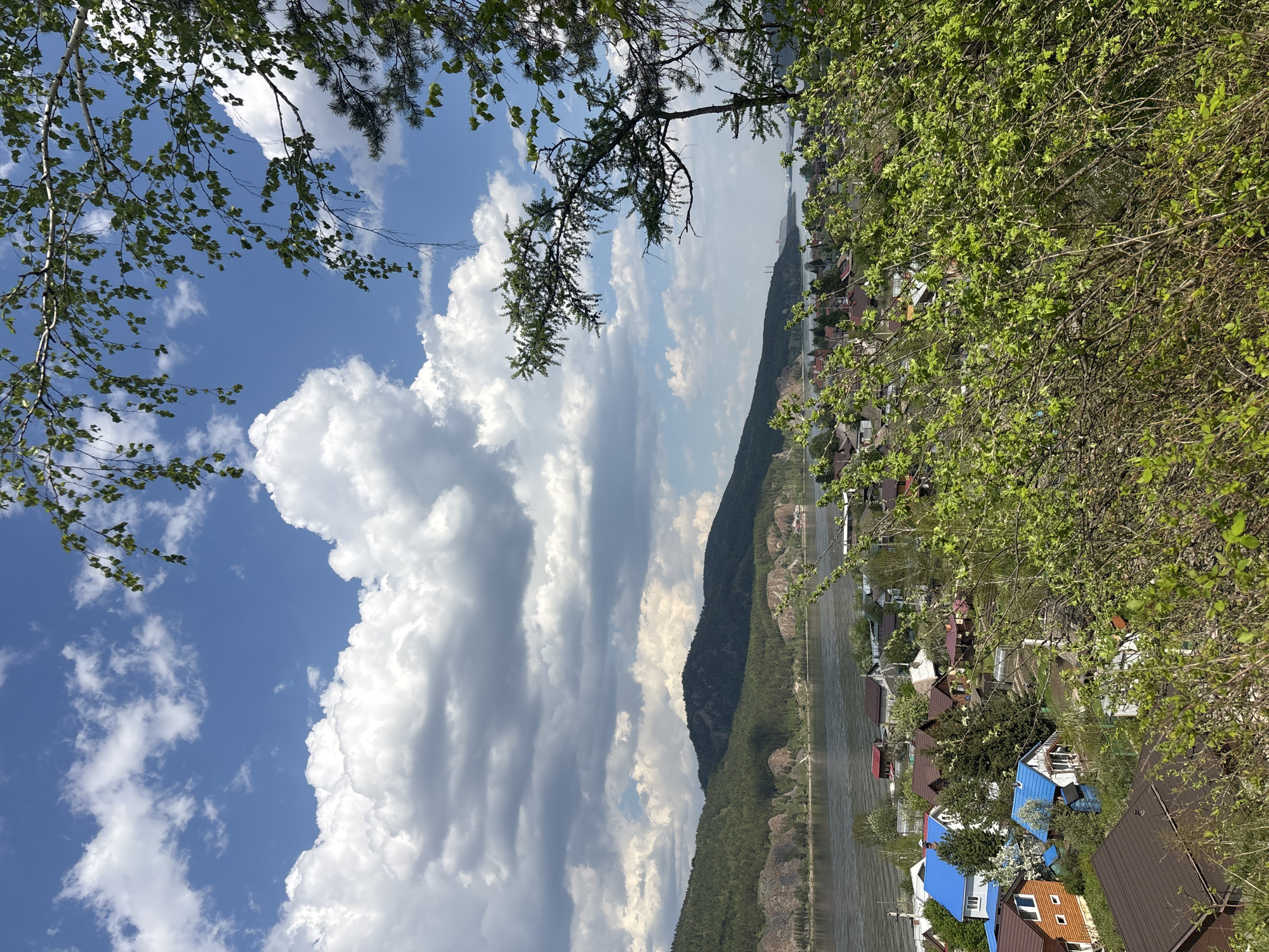The image depicts a landscape scene in Овсянка, Russia, with a combination of natural and built environments. In the foreground, there are branches of trees with fresh green leaves, partially framing the view. The midground features a river or lake, bordered by a wooded mountain range on one side and a settlement of houses on the other. The houses show a variety of roof colors, including blue and brown. In the background, there's a sky filled with white, puffy clouds interspersed with patches of blue. The overall scene conveys a sense of a rural or suburban area with a mixture of water, mountains, and small-scale residential architecture.
