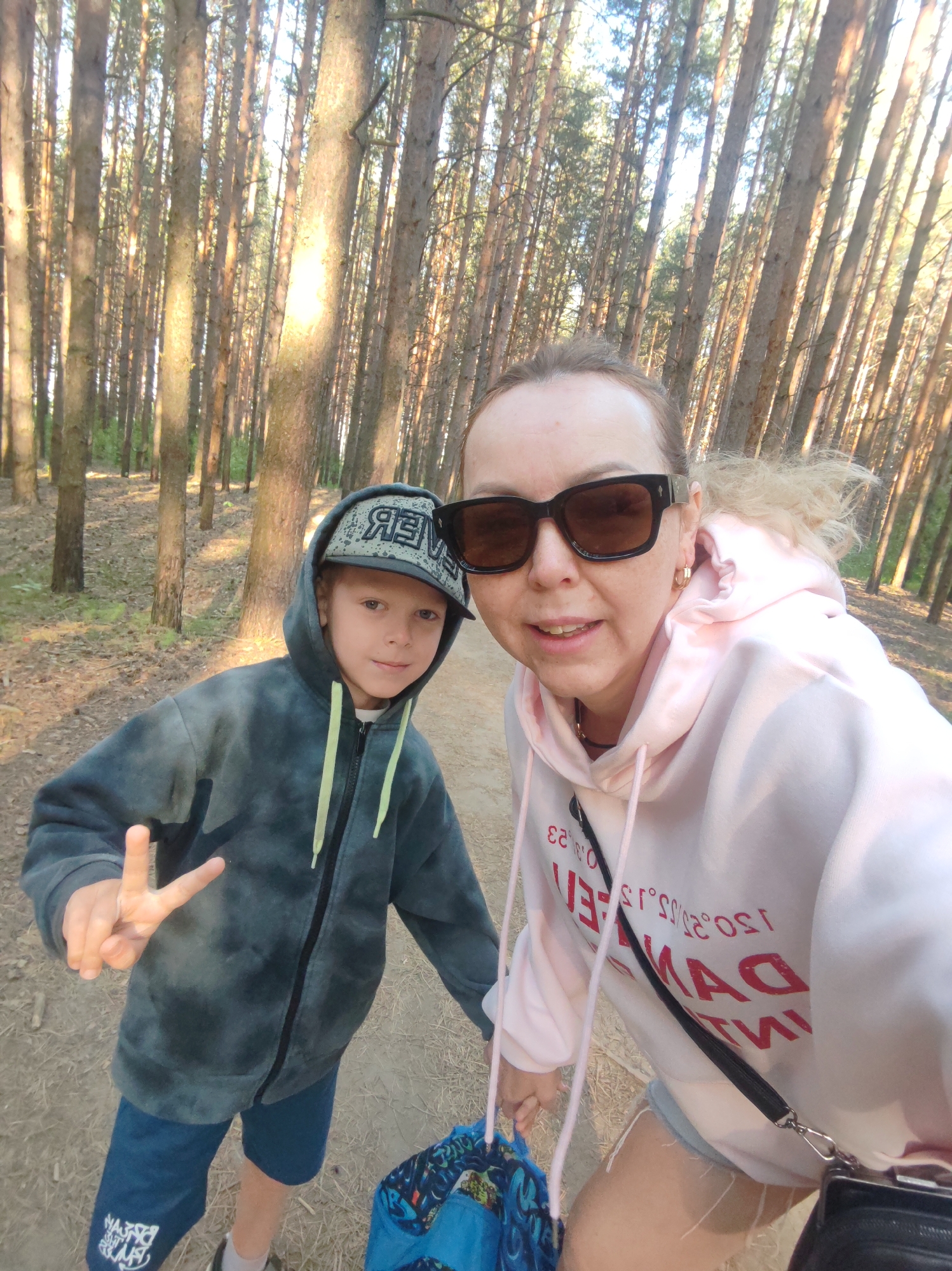 A selfie taken in a forest near Chaikovsky, Russia. A woman with sunglasses and a light pink hoodie stands next to a young boy in a hoodie and baseball cap. The boy is holding up a peace sign. The forest is densely populated with tall, thin trees. A dirt path is visible in the foreground. The woman has a black purse or bag slung over her shoulder.