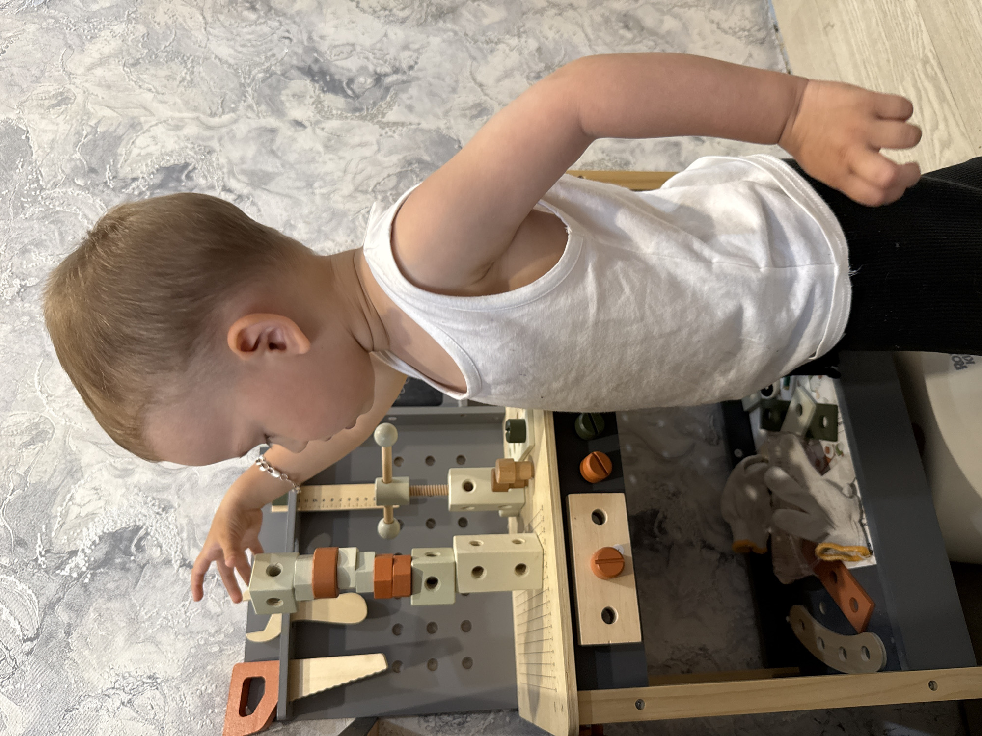 A young child wearing a white tank top and black pants is playing with a wooden toy workbench. The child has short blonde hair and is wearing a silver bracelet on their left wrist. The workbench is light gray and brown, with various wooden tools and shapes attached to it. The workbench includes a vise, a ruler, a saw, and several wooden blocks and screws. A pair of gloves and additional wooden pieces are on the bottom shelf of the workbench. The background shows a textured gray and white wall. The scene appears to be indoors. There is no verifiable information to confirm the location in the city of Rusanovo, Russia.
