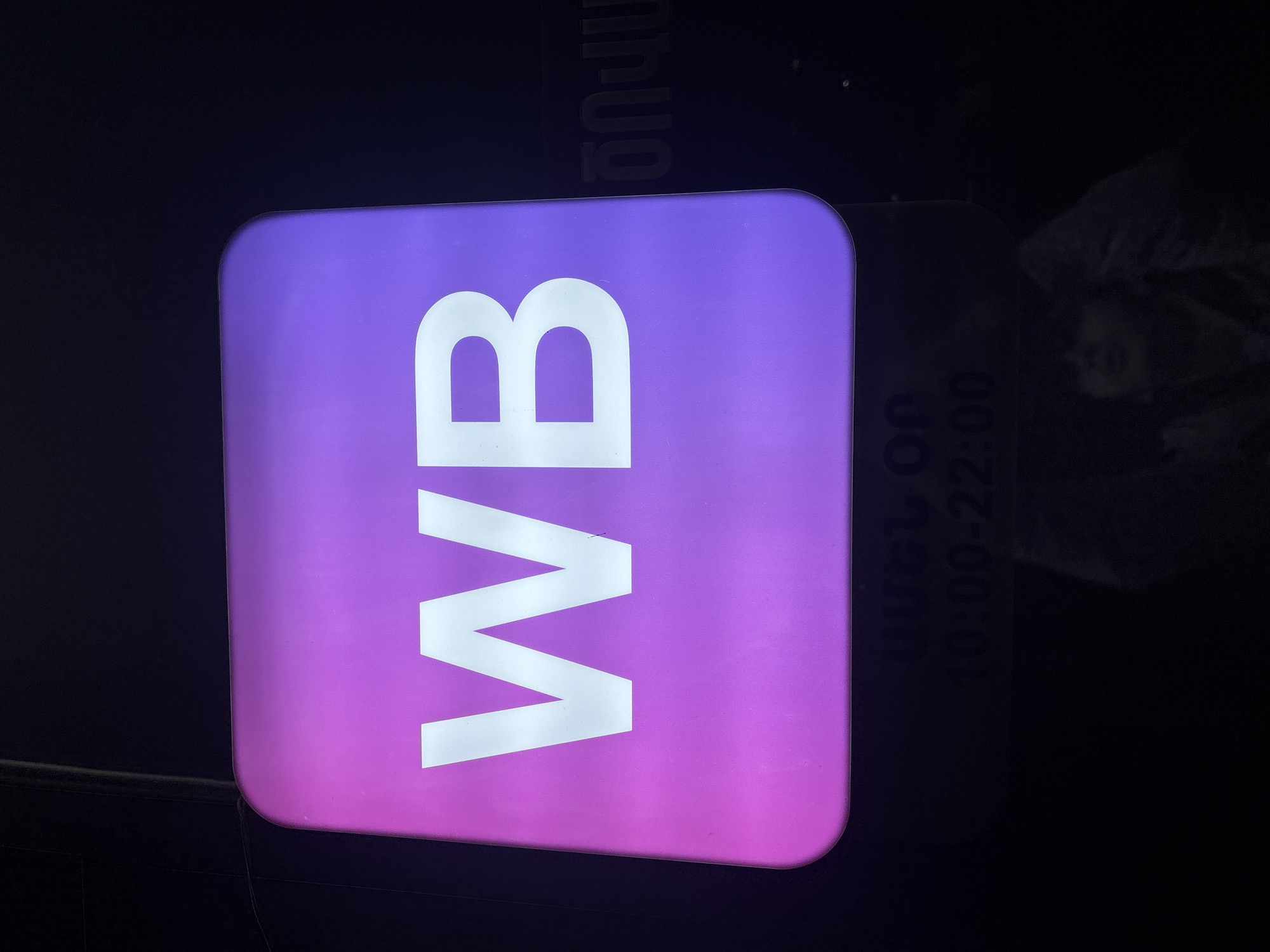 Here's a description of the image:
The image depicts a sign with the letters "WB" in white on a square purple-to-pink gradient background. The sign appears to be illuminated. To the right of the sign, barely visible, is additional text that includes the time "10:00-22:00", potentially indicating opening hours. The sign is located in Yerevan, Armenia.
