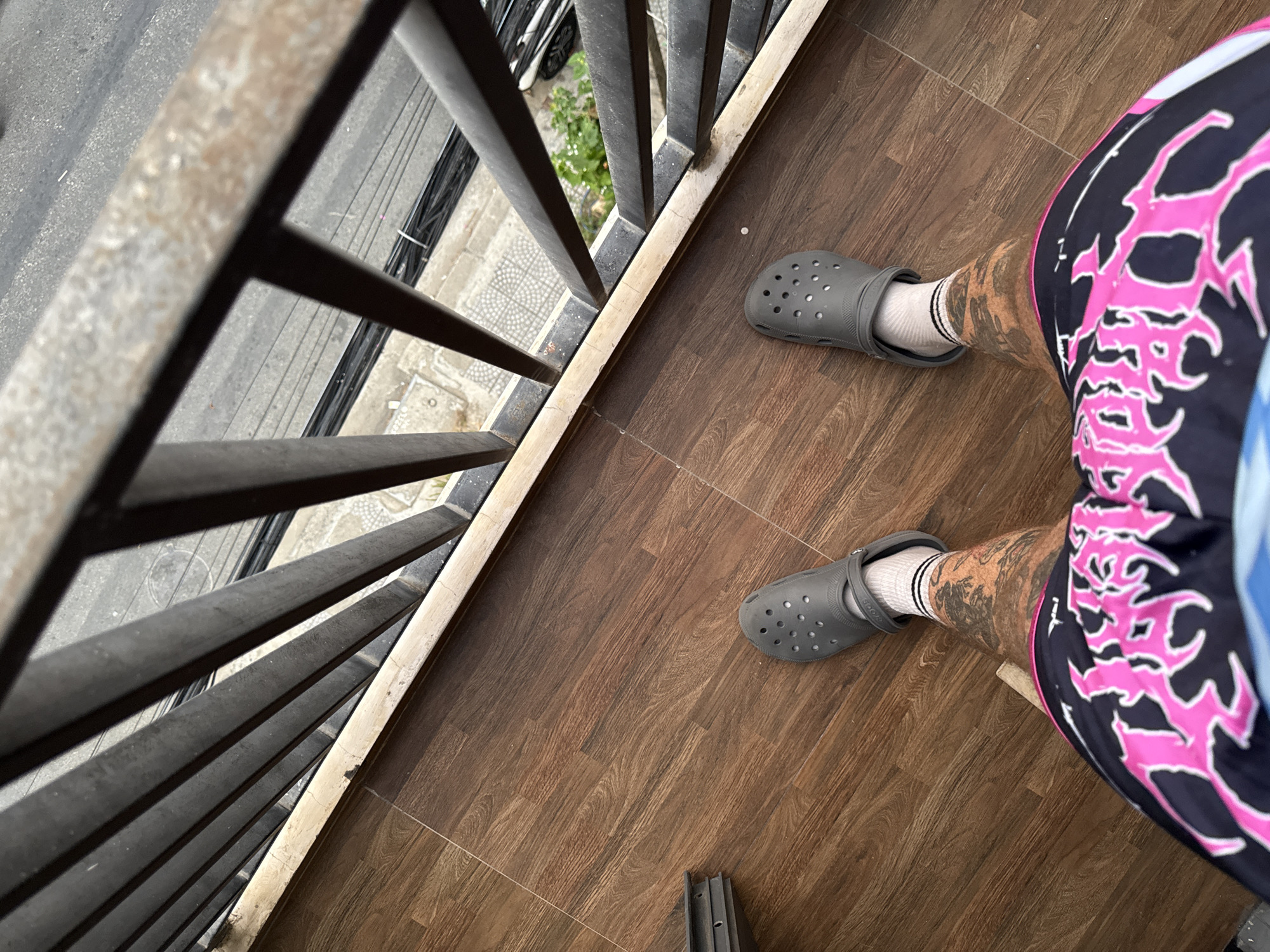 This overhead image captures a person's lower body standing on a tiled balcony or elevated platform, overlooking a street scene below. The individual wears dark grey Crocs clogs with white socks that have black stripes near the cuff. The left leg displays extensive tattooing. The person's shorts are black, featuring prominent, jagged bright pink and white graphic designs, which appear to be stylized text or symbols.
The floor is covered with brown ceramic tiles designed to mimic wood planks. A dark metal railing with vertical bars, showing some signs of wear and discoloration, separates the balcony from the view below.
Beyond the railing, a street or alleyway is visible, characterized by a grey asphalt or concrete surface. Bundled electrical cables are noticeable, running across the upper part of the street view. Geometric patterned sidewalk tiles and some green foliage from trees or bushes are also discernible. The scene is illuminated by natural daylight. The urban landscape details, including the street infrastructure and building materials, are consistent with a setting in Đà Nẵng, Vietnam. The word "CROCS" is visible on the strap of the left shoe.