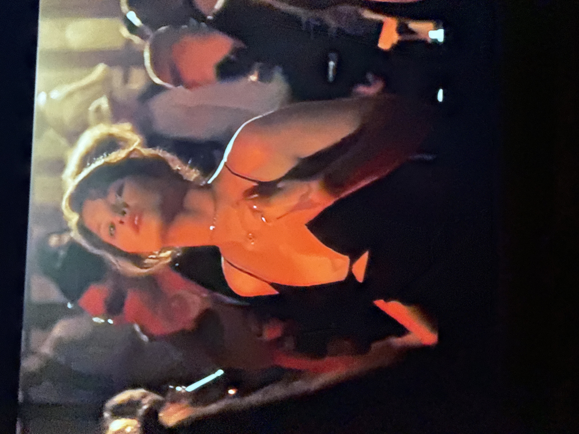 A woman with her hair in a messy bun and wearing a black dress with spaghetti straps is illuminated by a warm light source, possibly a spotlight. She is in the midst of a crowded environment, with blurred figures surrounding her. The environment is dark overall. The location and specific details of the city of Mytishchi, Russia, cannot be determined from this image.